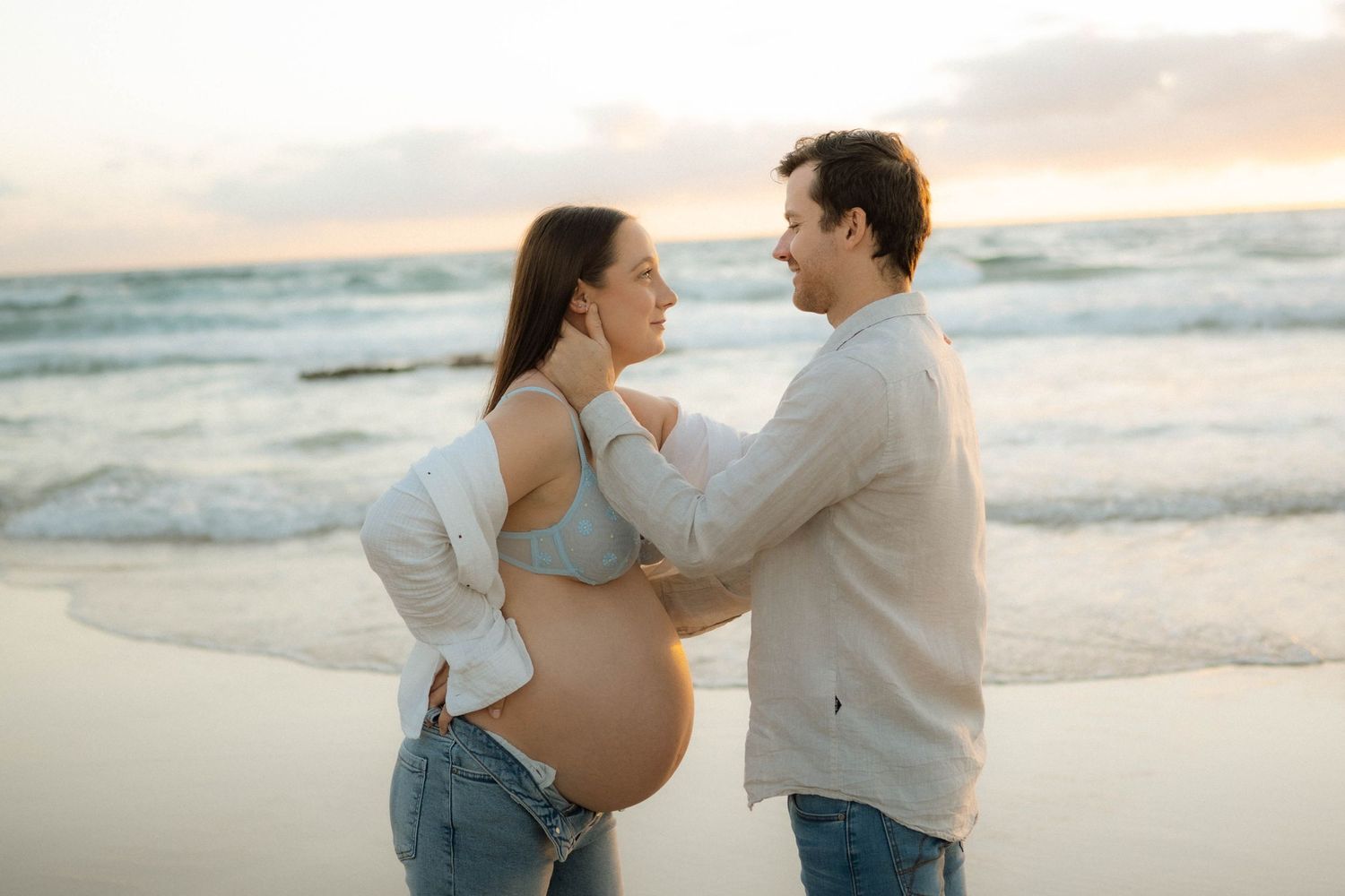 Smiling couple embracing on beach at sunset, showcasing pregnant belly in casual attire.
