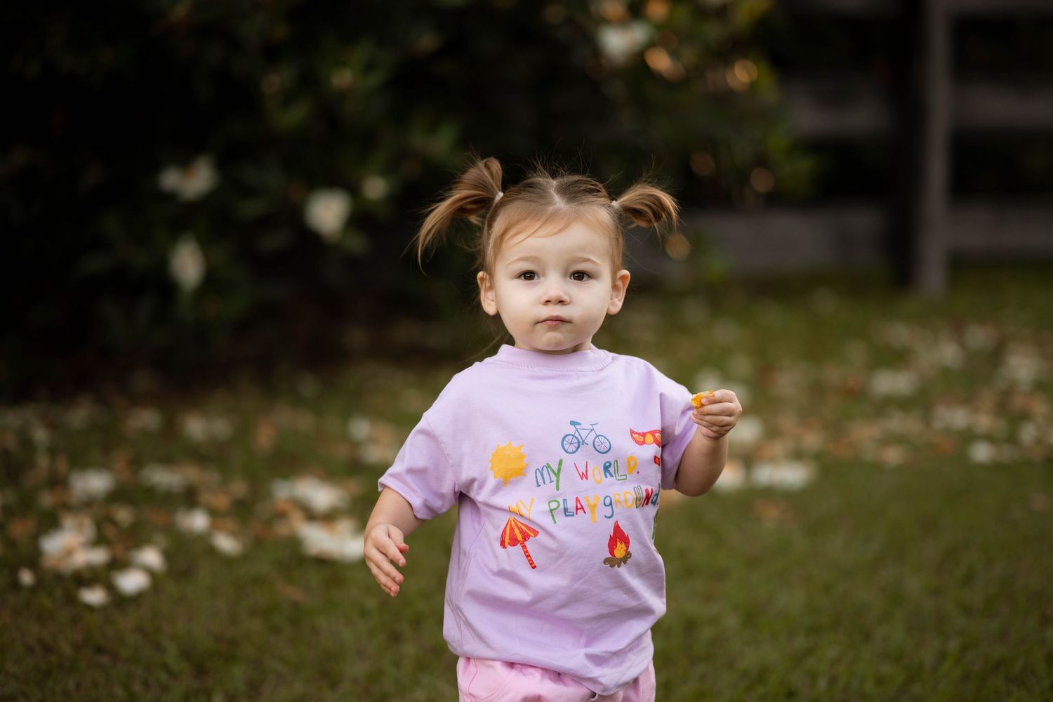 Toddler girl with pigtails wearing a lavender graphic tee runs outdoors on green grass.