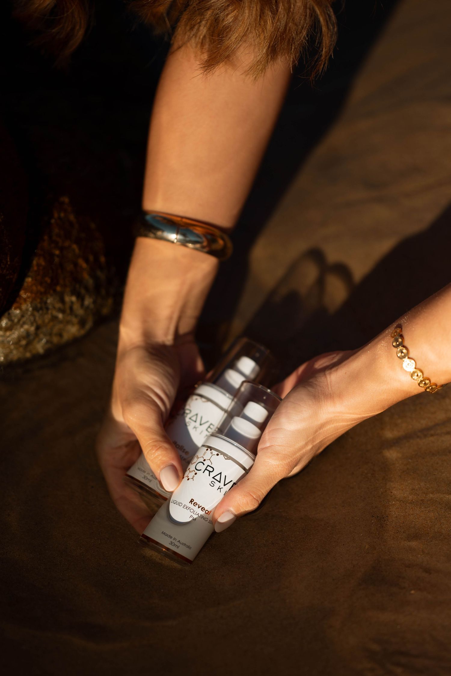 Hands holding Crave skincare serum and cream products in warm sunlight.