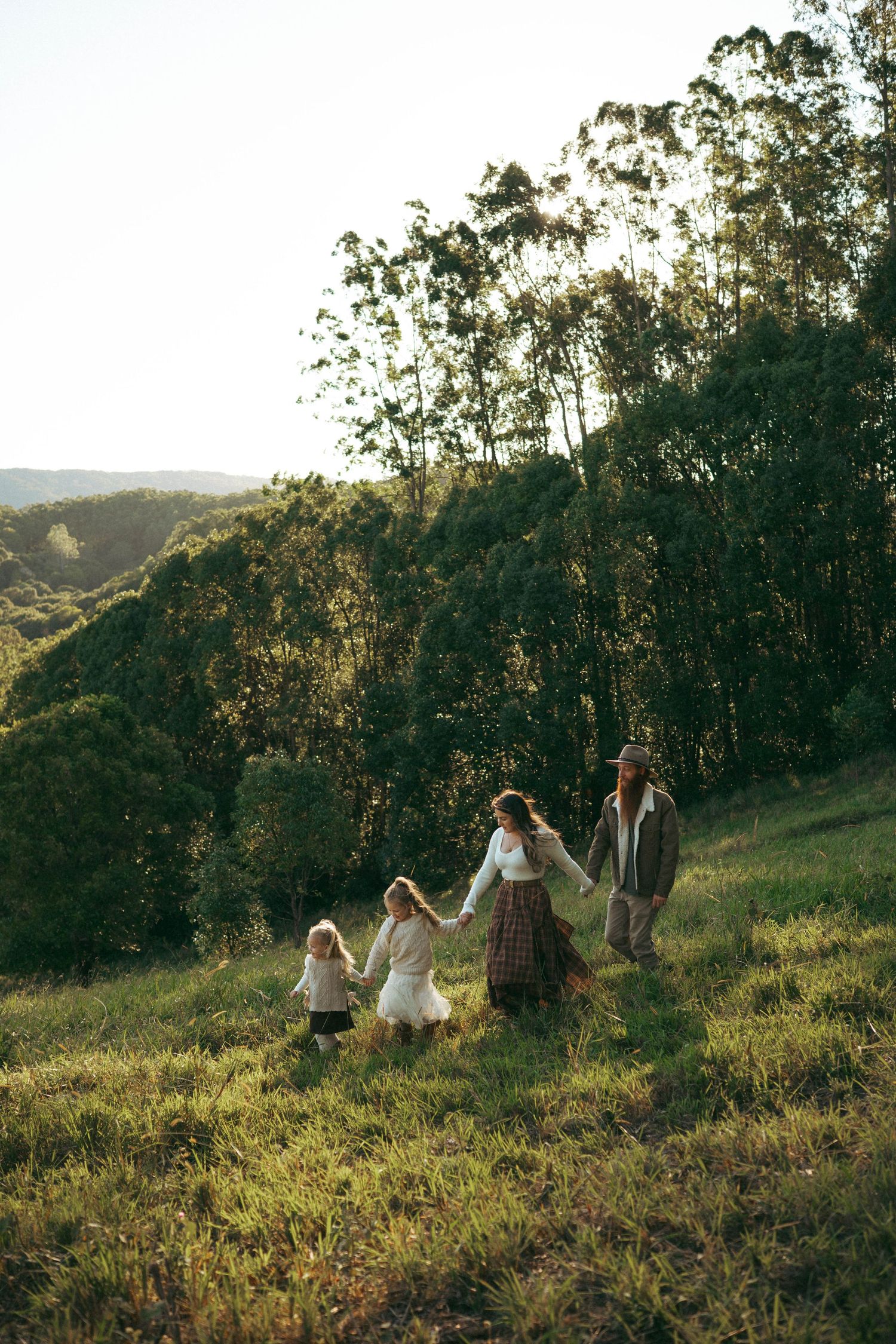 Family of four holding hands walking through lush green hillside meadow at golden hour sunset.