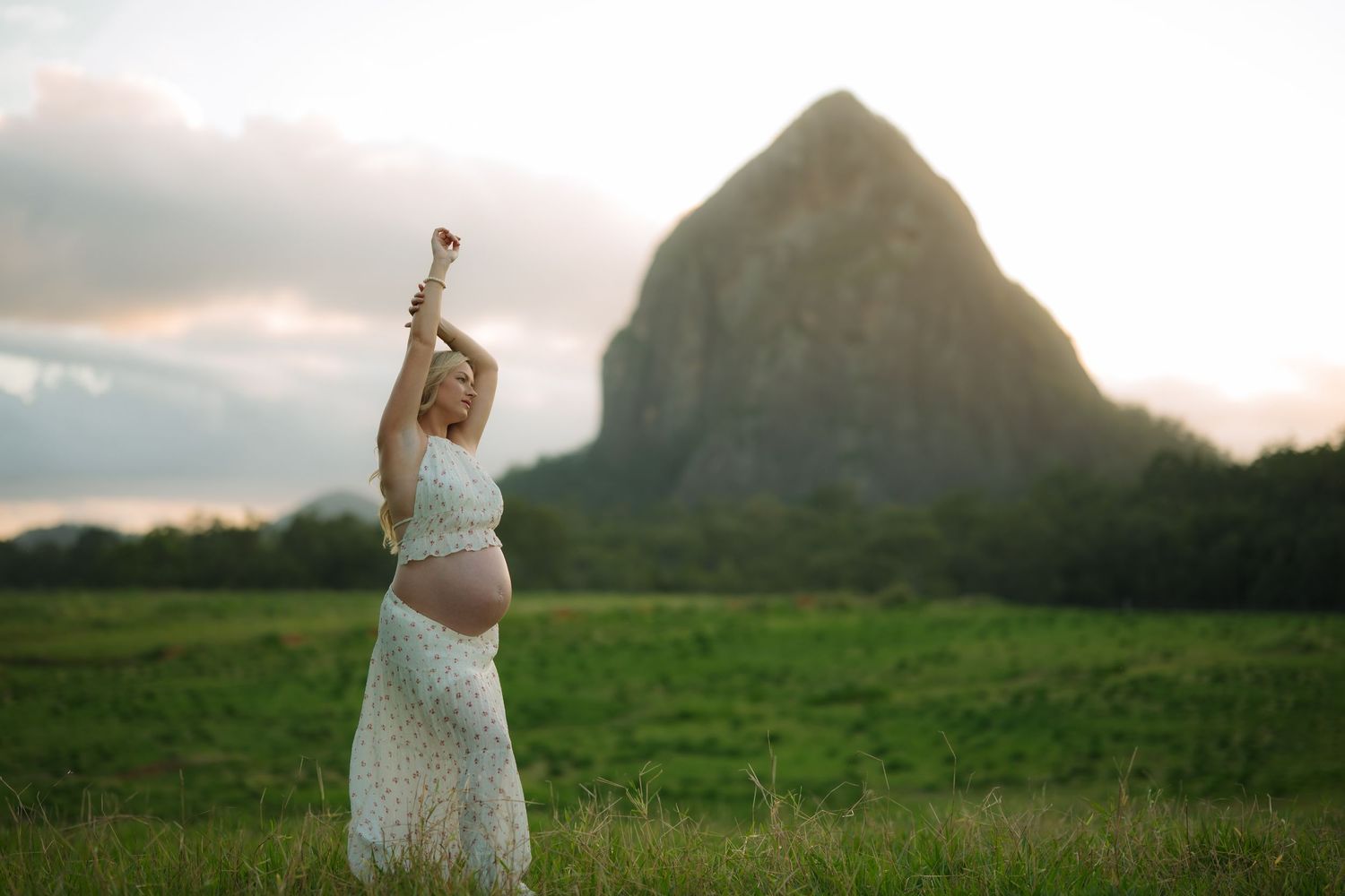 Pregnant woman in white floral dress raises arm joyfully in green field with large rock formation at sunset.