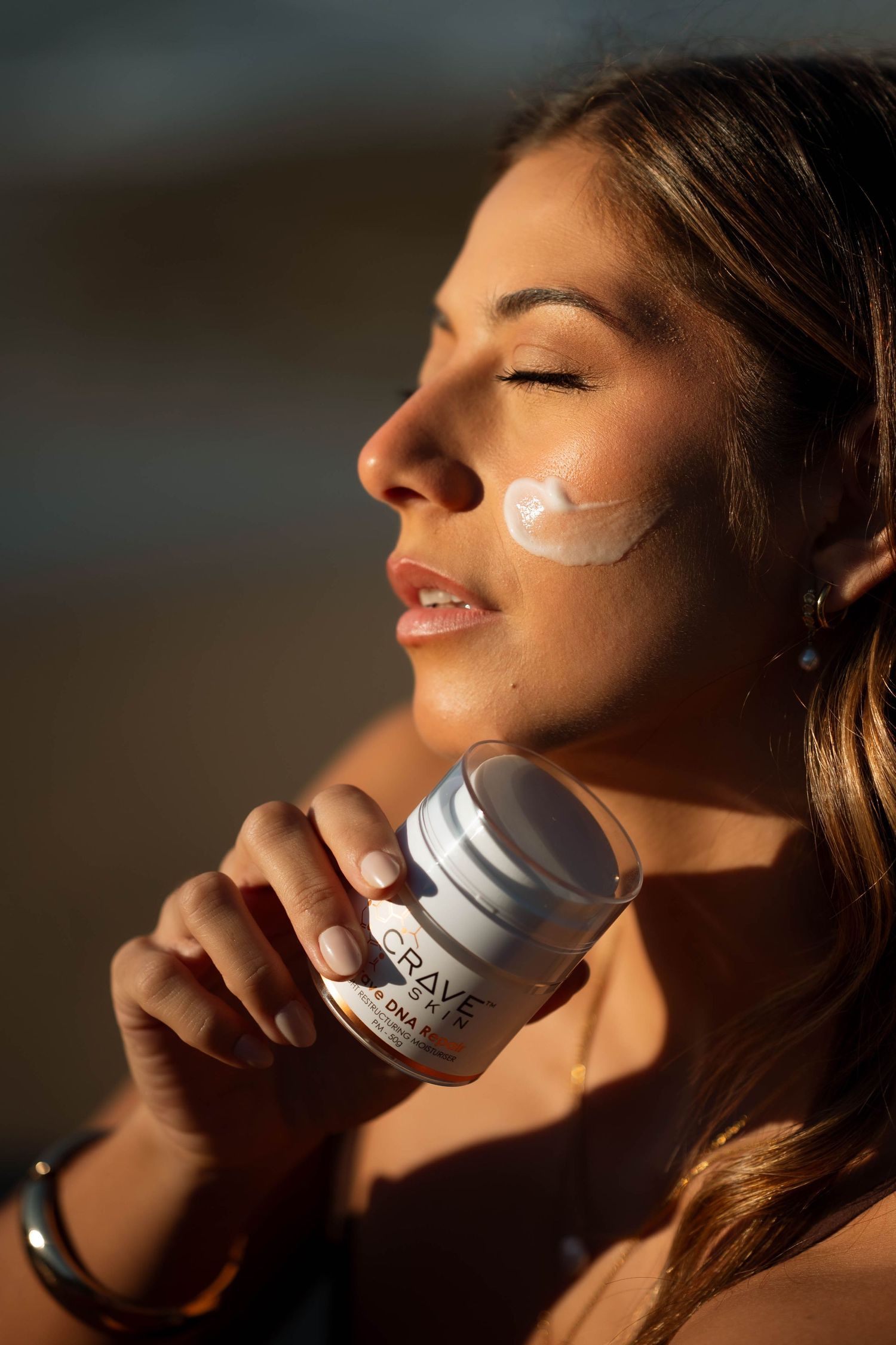 Woman applying Crave Skin DNA moisturizer to her cheek in warm sunlight.
