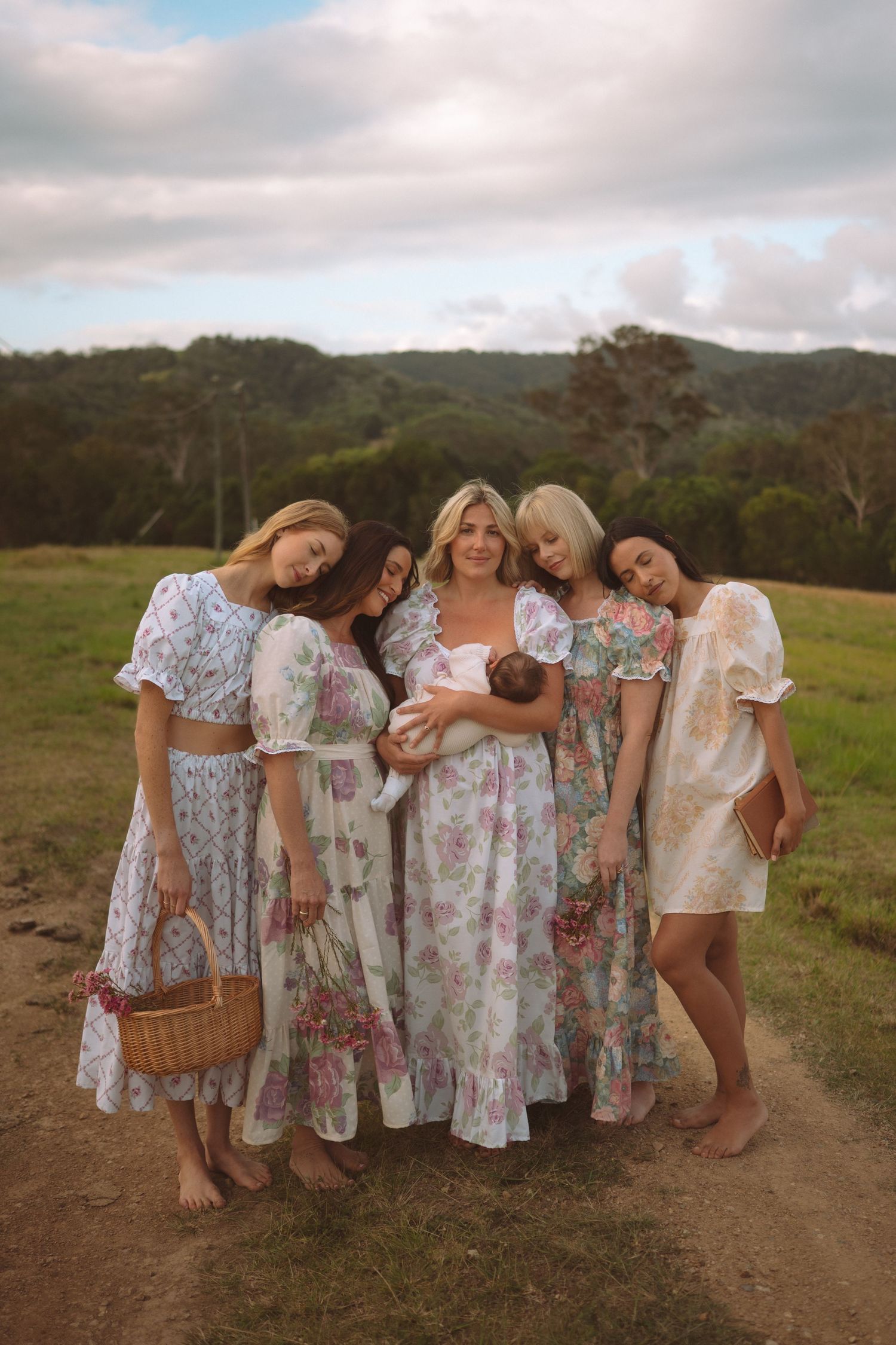 Five women in floral dresses gather outdoors on a dirt path, one holding a newborn baby, with green hills behind.