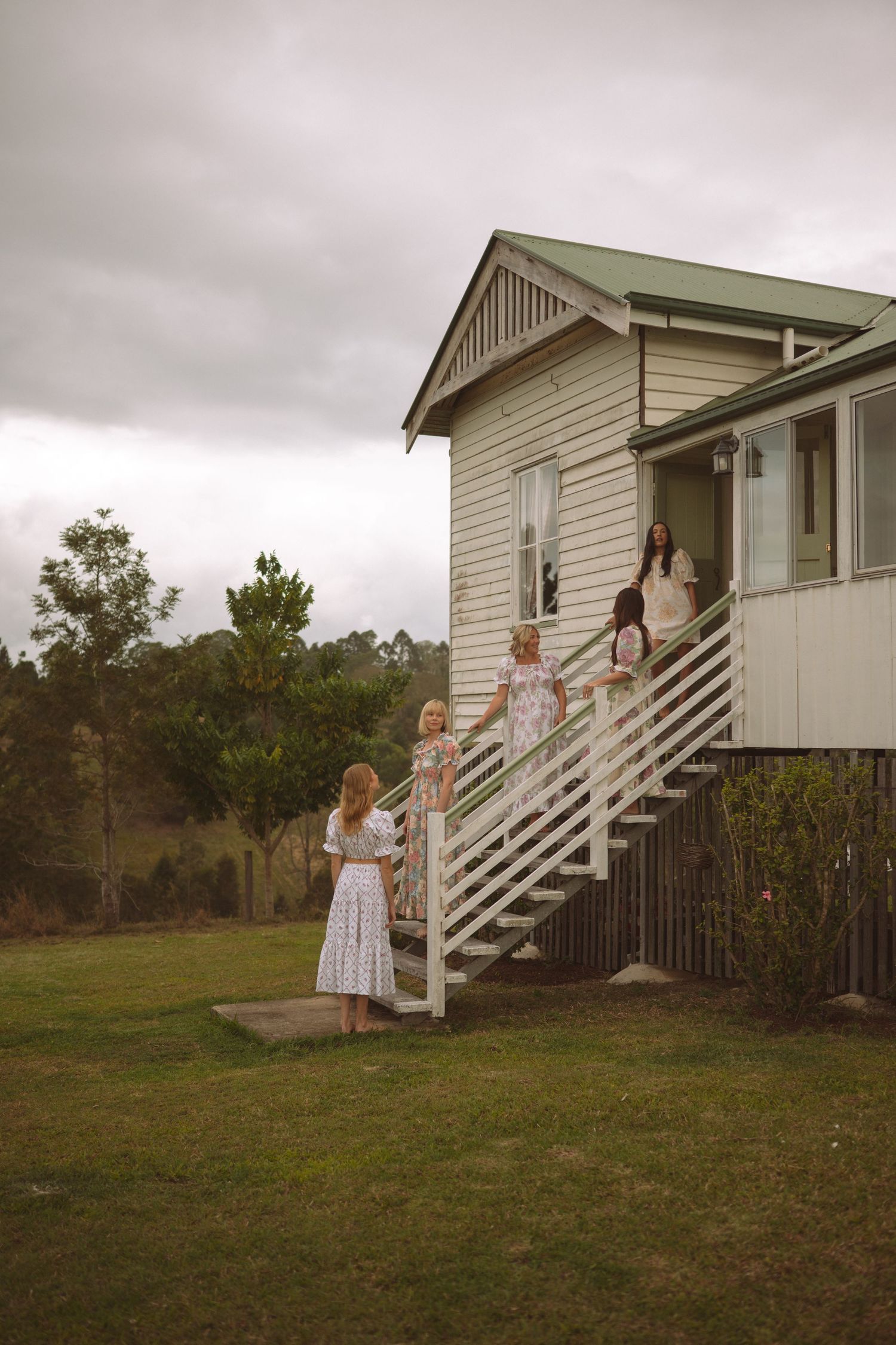 Women in white dresses descending porch stairs of a rustic white weatherboard home on a cloudy day.