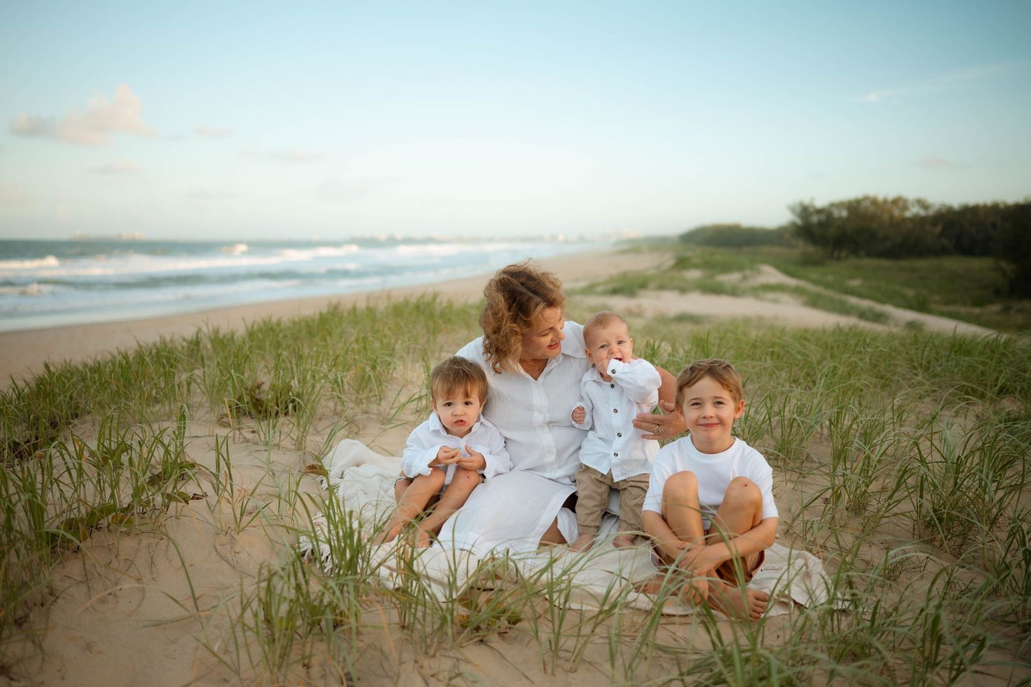 Mother with three young boys in white shirts sitting on beach sand dunes at sunset.