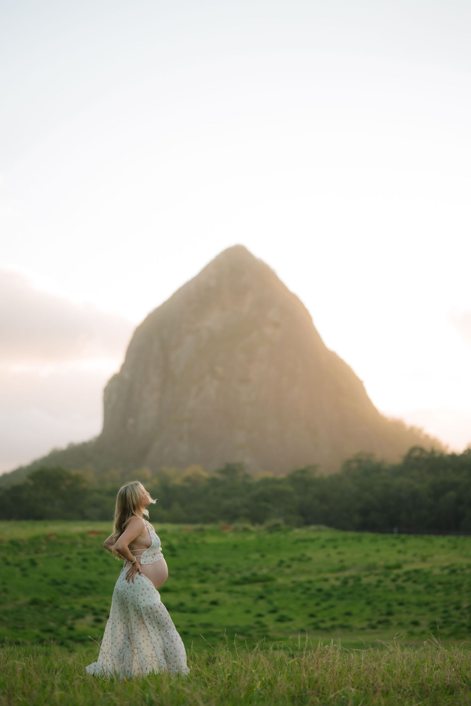 Pregnant woman in flowing white dress stands gazing up at a sunlit rocky peak in a lush green meadow.