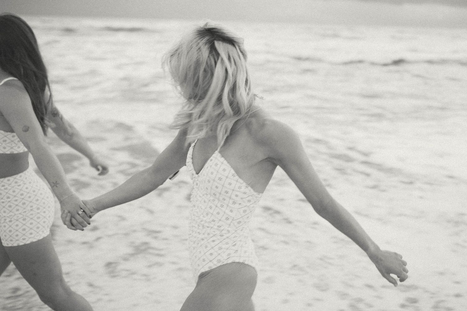 Two women holding hands running along the beach shore in white swimsuits in black and white photo.