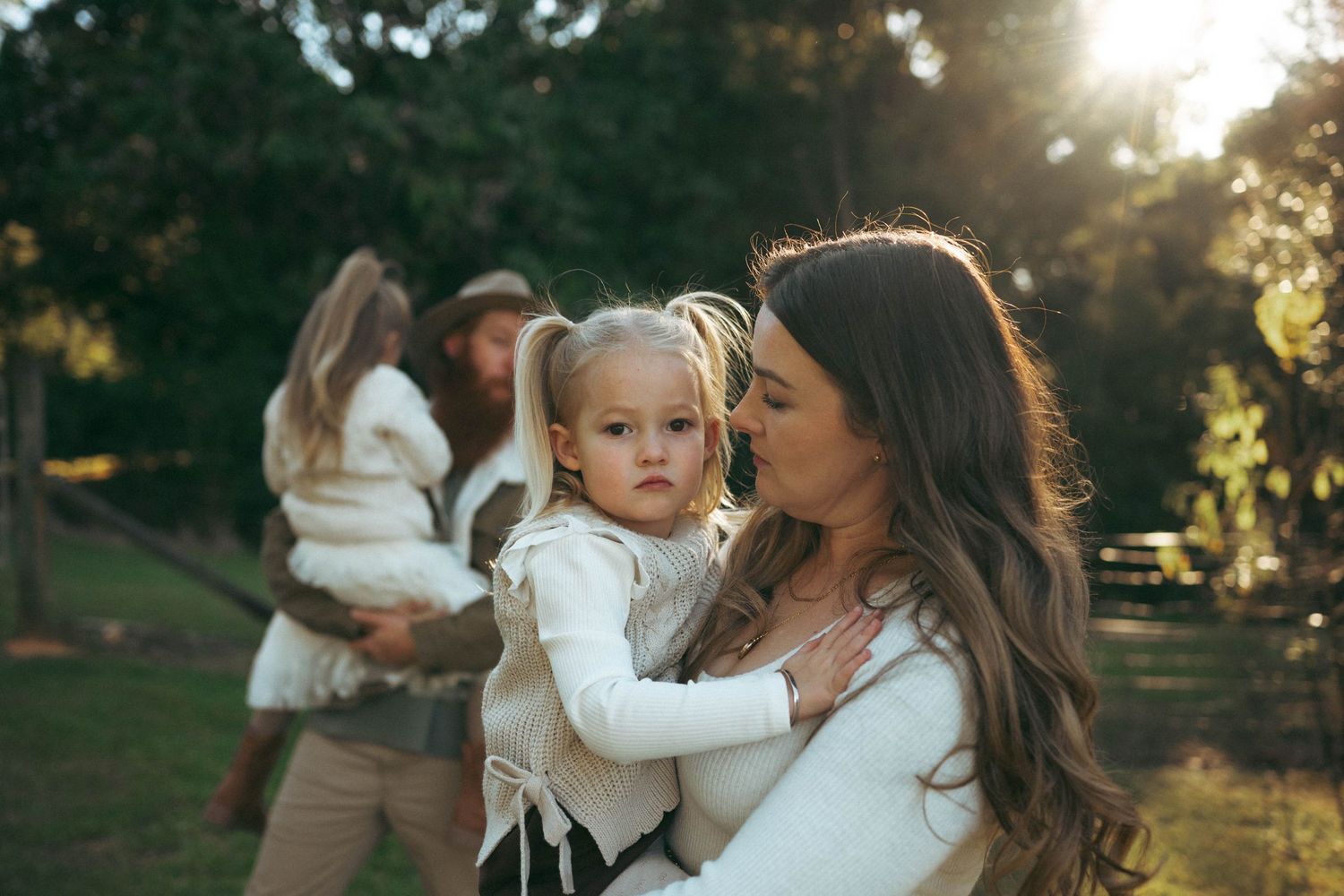 A mother kisses her toddler daughter outdoors in a sunlit park, with a family in the background.