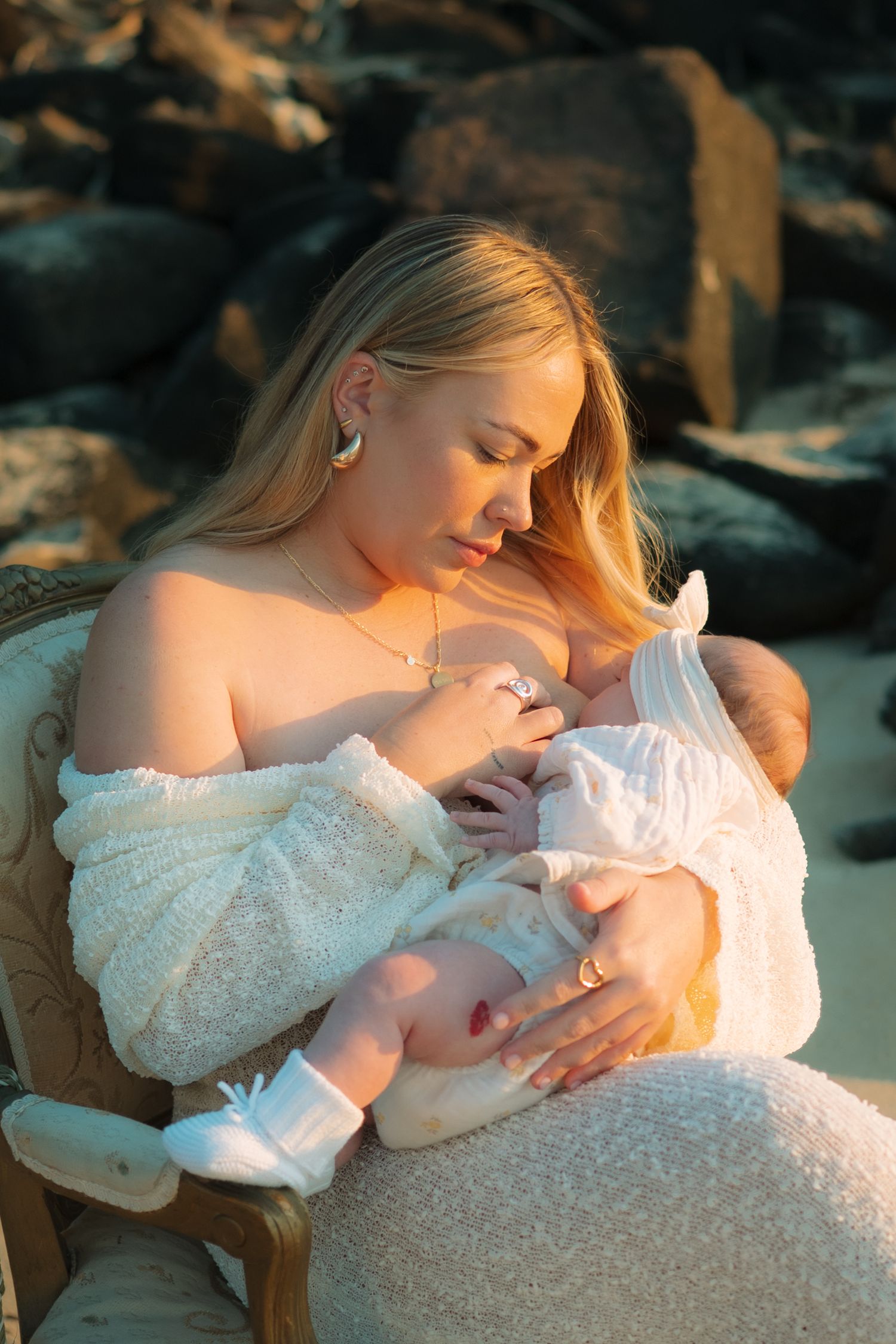 Blonde mother in white off-shoulder dress breastfeeding newborn baby outdoors in golden sunset light.