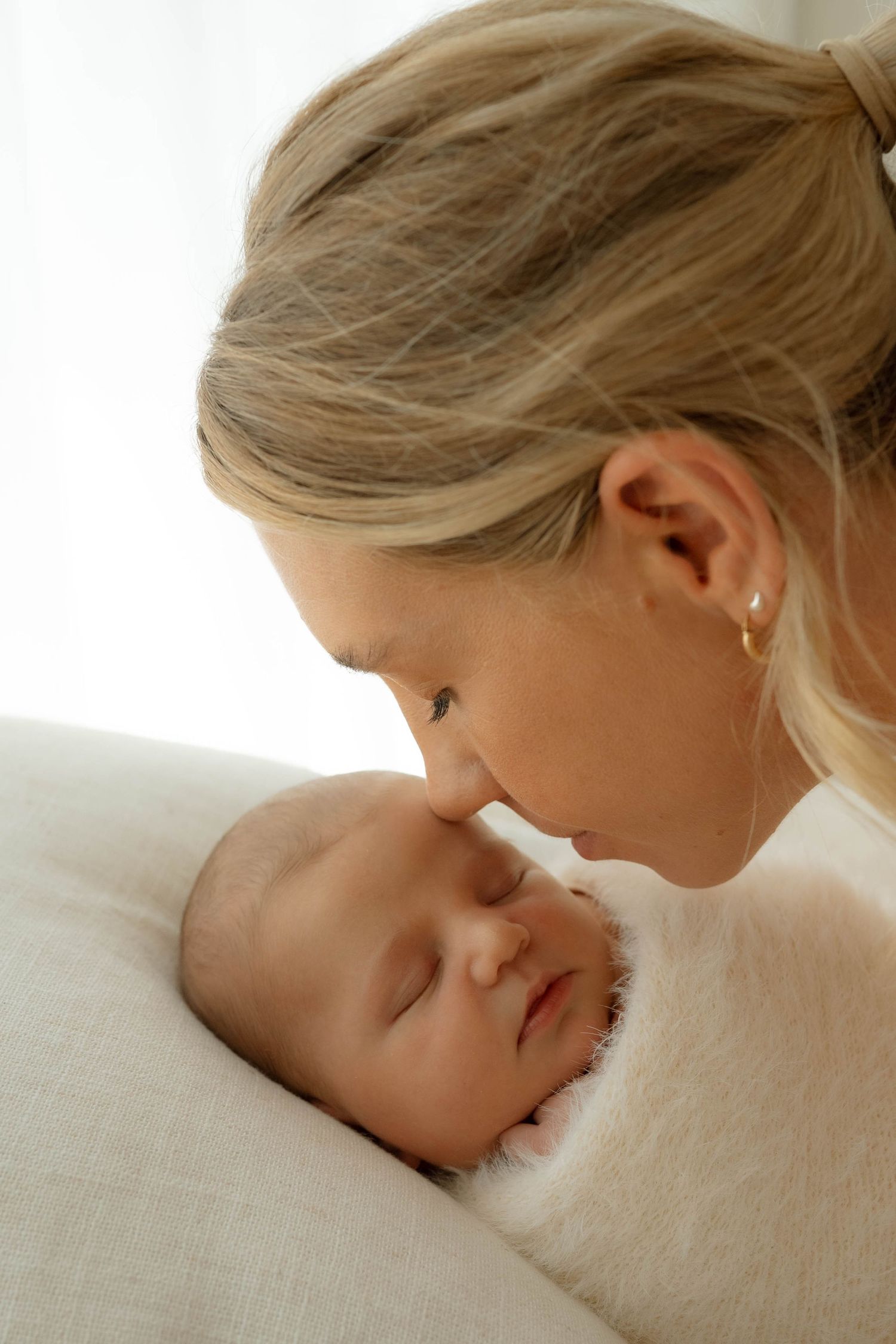 Tender moment of a blonde mother gently touching noses with her sleeping newborn baby.