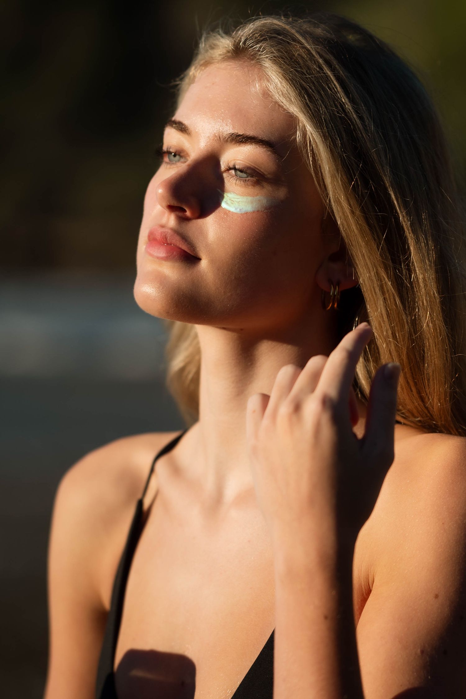 Young woman applying sunscreen to her face outdoors in warm sunlight, wearing a black bikini top.
