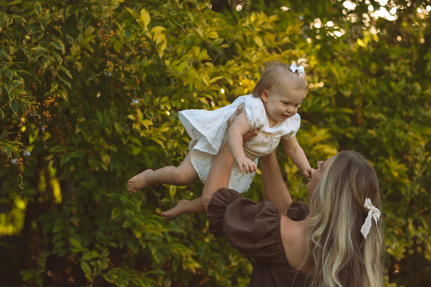 Mother lifting smiling baby girl in white dress among green trees.