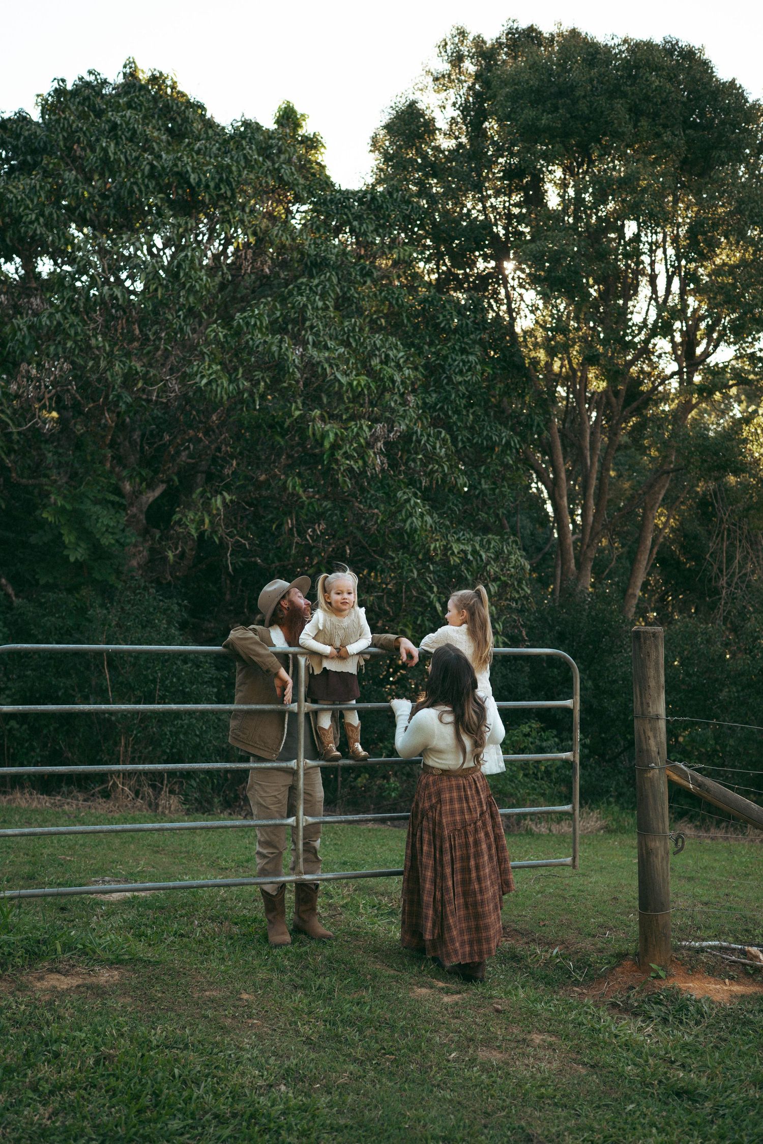 Family with baby at farm gate surrounded by lush green trees at golden hour.