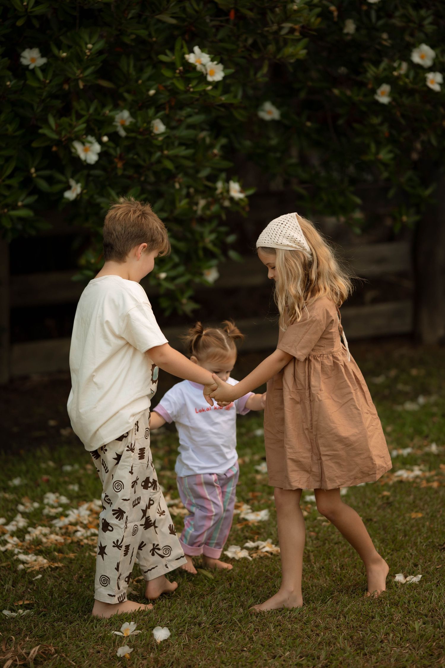 Three barefoot children holding hands and playing together on green grass near a flowering bush outdoors.