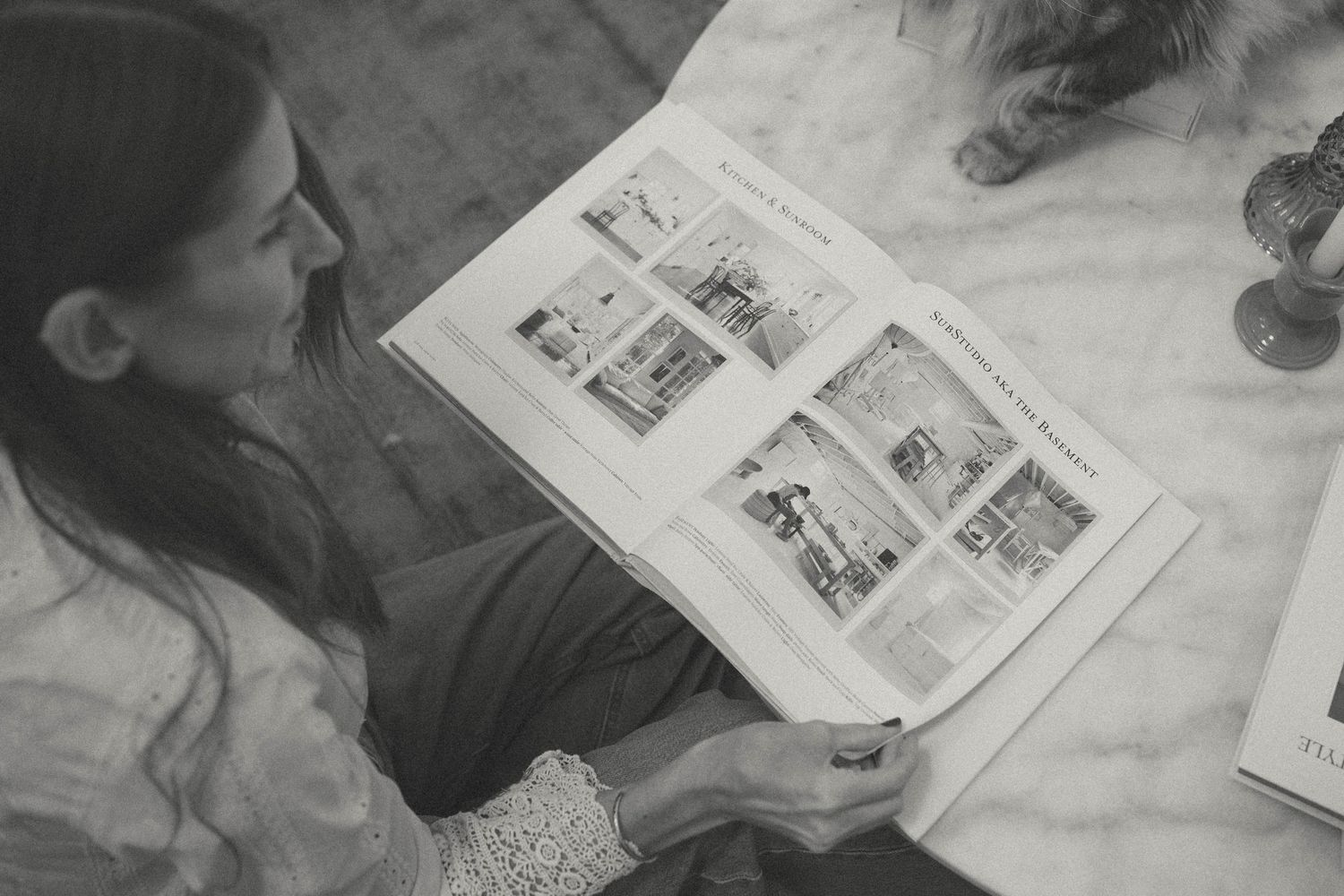 Black and white photo of a woman reading an interior design magazine at a marble table.