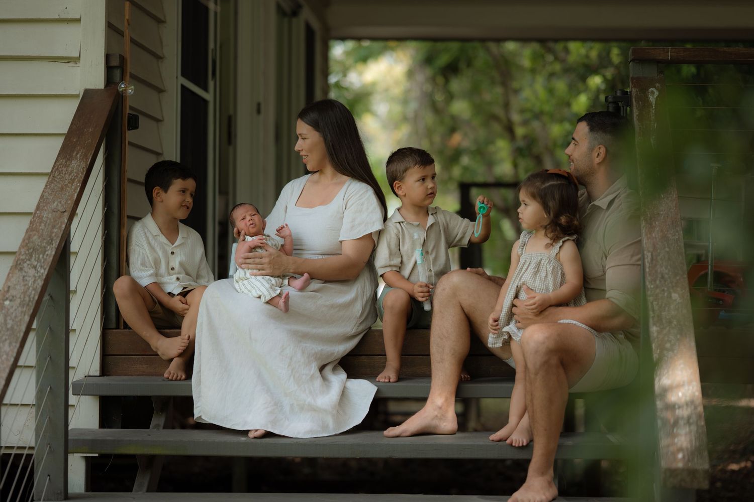Family portrait on wooden porch steps, mother in white dress holding newborn with three children and father.