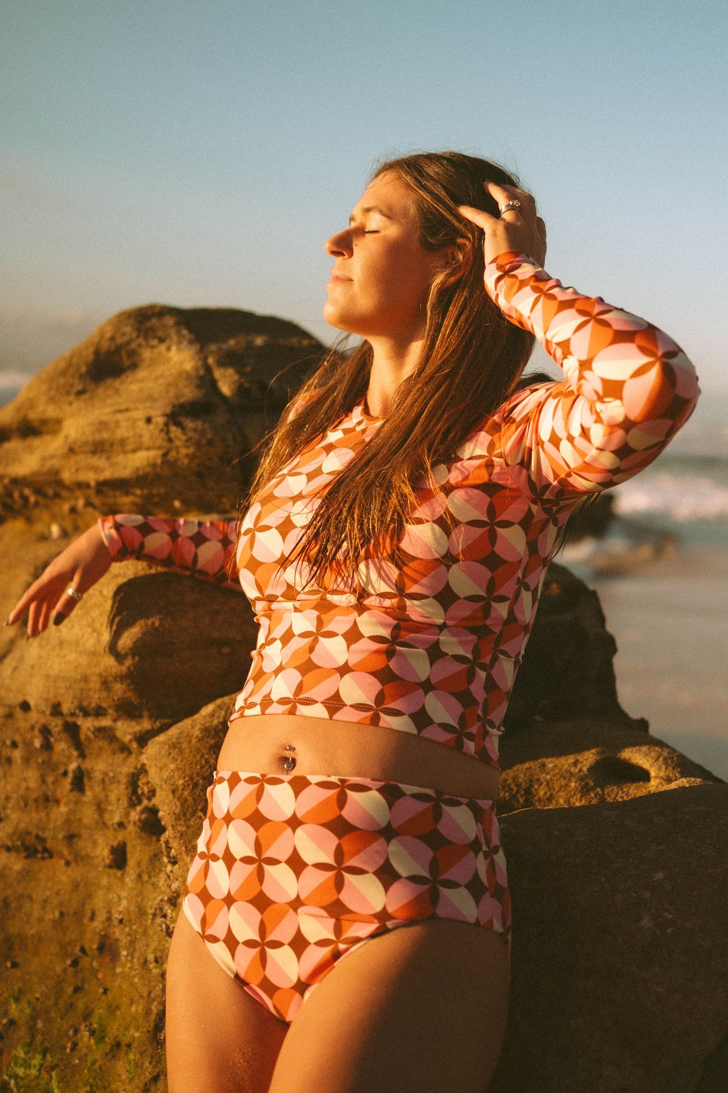 Woman in orange geometric print long-sleeve crop top and high-waist bikini bottom posing by beach rocks.