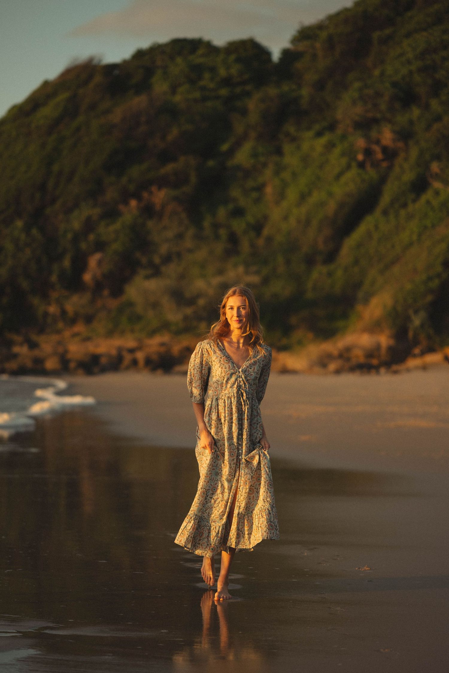 Woman in floral maxi dress walking barefoot on a sandy beach at golden hour with lush green cliffs behind.