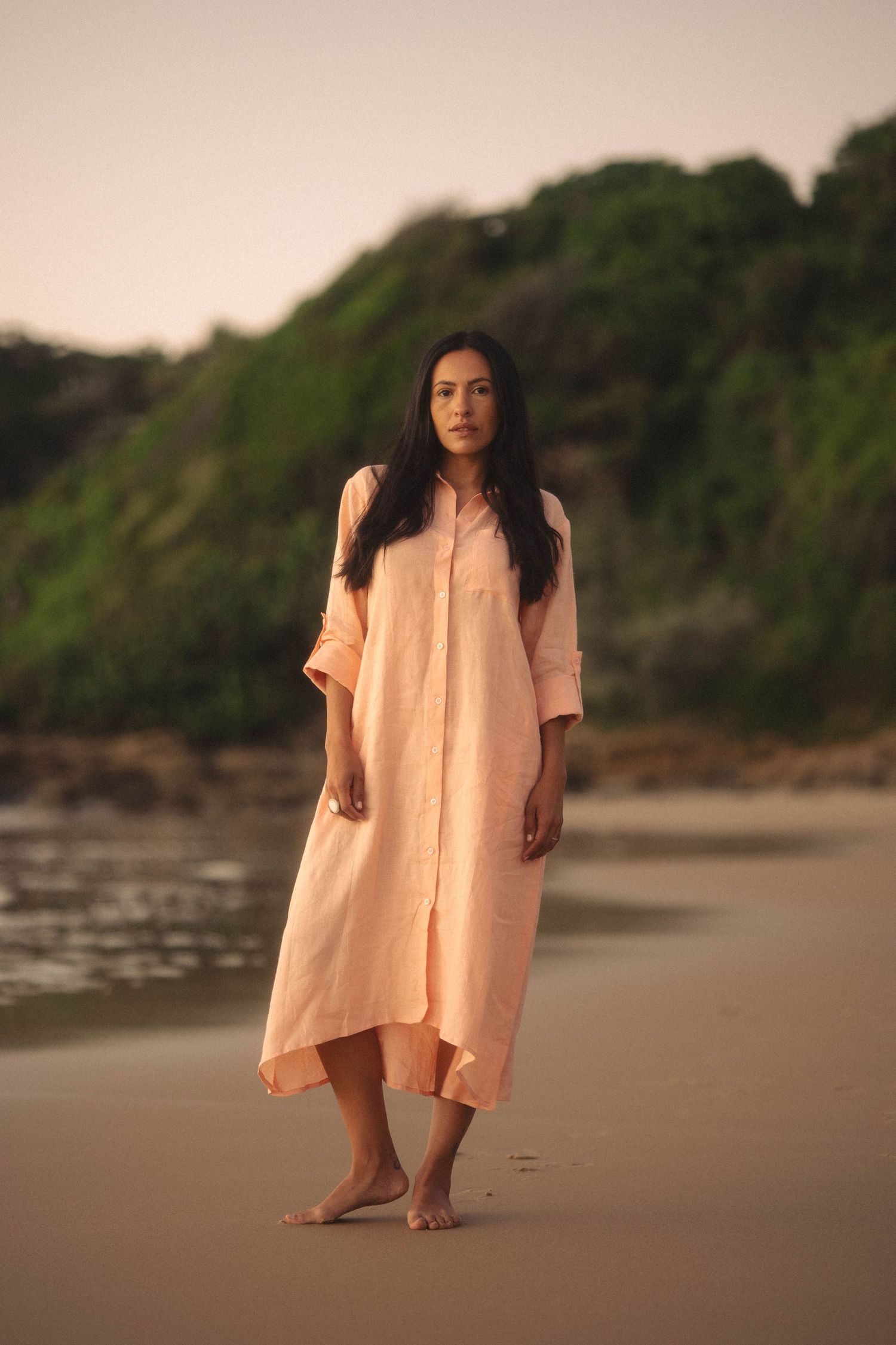 Barefoot woman wearing long peach linen dress walking on beach with green cliffs behind.
