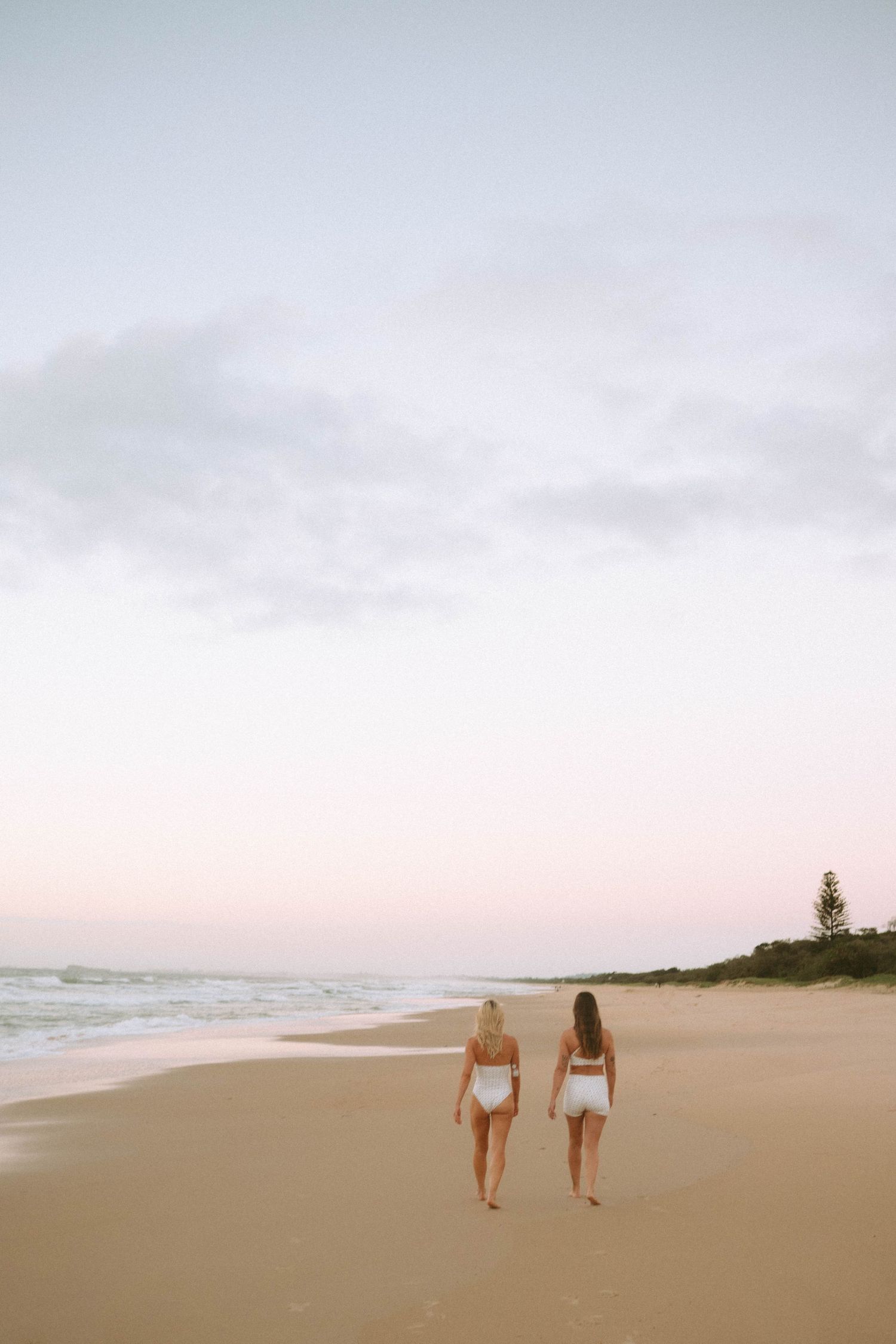 Two women in white bikinis walking along a sandy beach at pastel sunset.