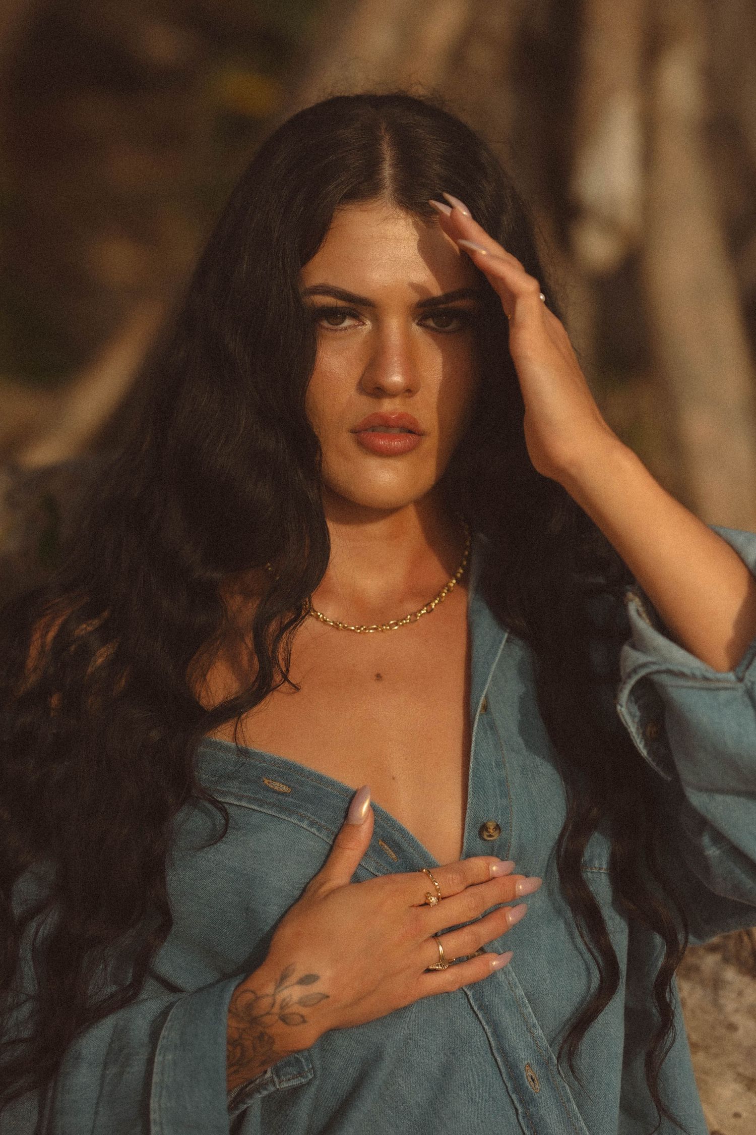 Dark-haired woman in open denim jacket wearing gold chain necklace and rings outdoors.