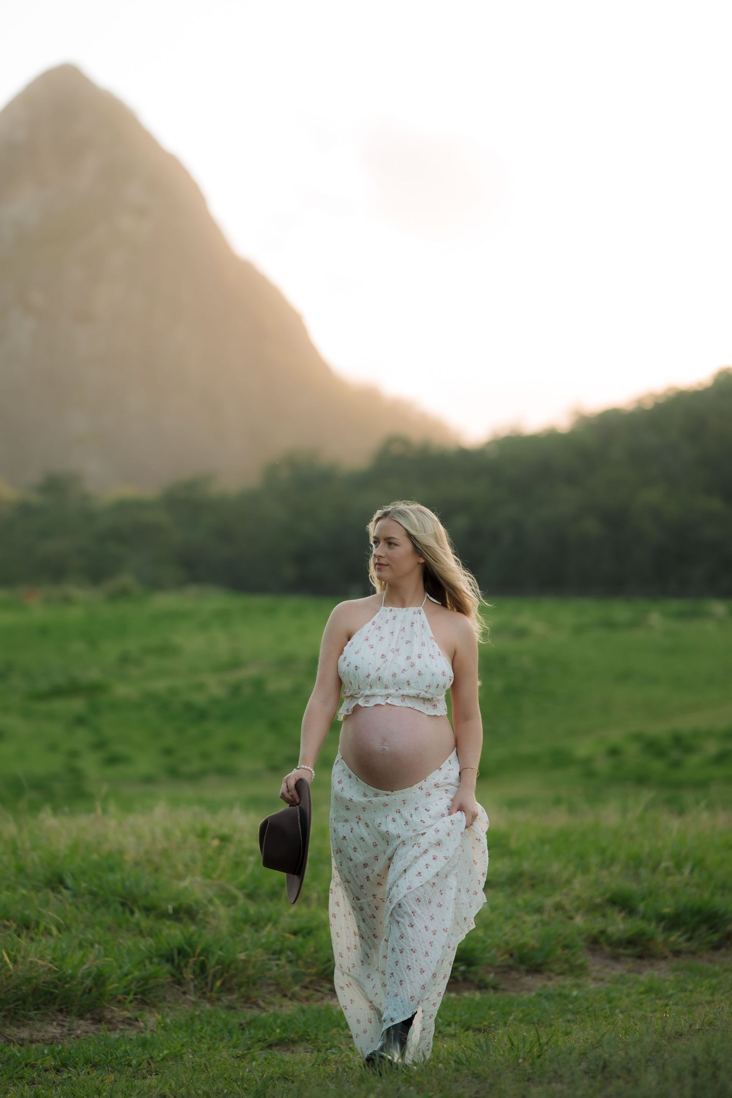 Pregnant blonde woman in floral halter maxi dress walking in green field at sunset.