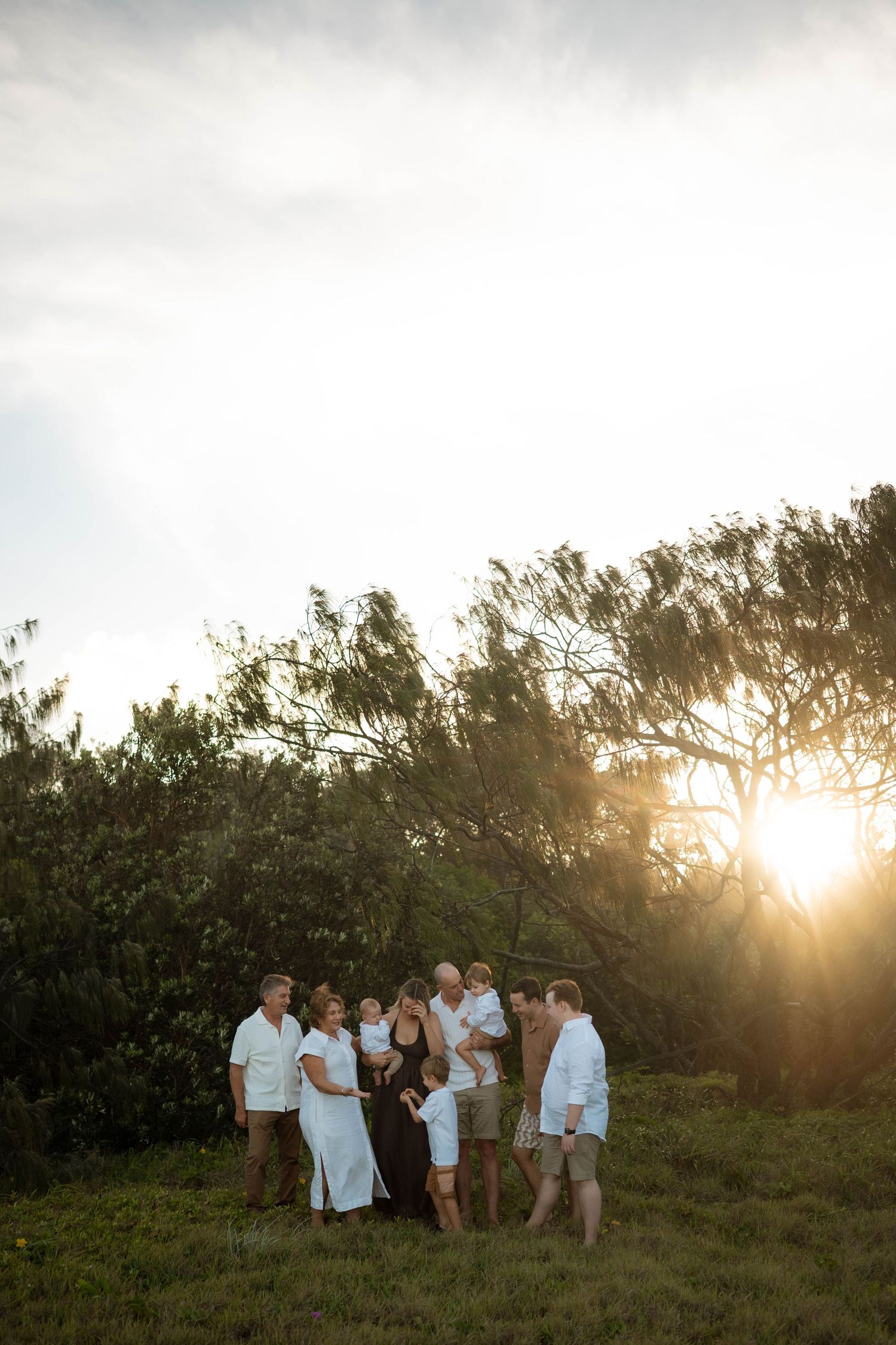 Large multigenerational family standing together in a sunlit field with dramatic golden sunset behind trees.