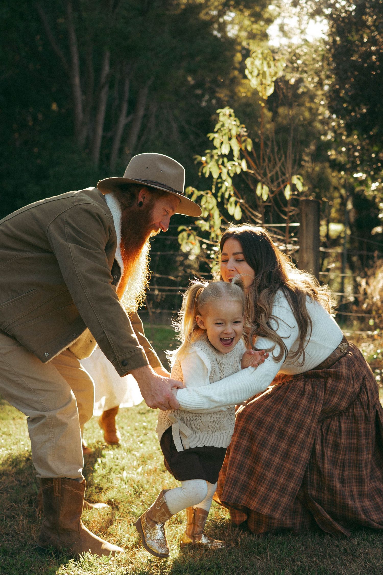 A bearded father in a hat and mother in plaid skirt laugh with their young daughter outdoors in golden sunlight.
