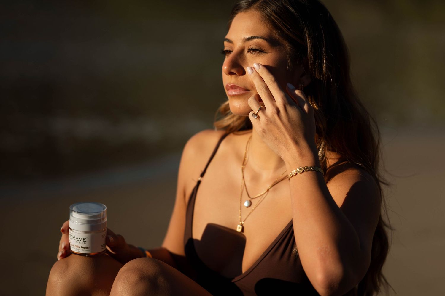 Woman applying skincare cream from a jar while wearing gold jewelry and a black top in warm sunlight.