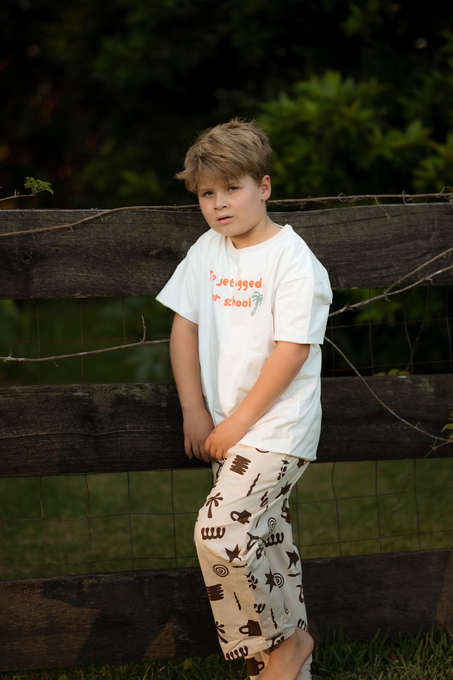 Young boy in white graphic tee and patterned shorts leaning against a rustic wooden fence outdoors.