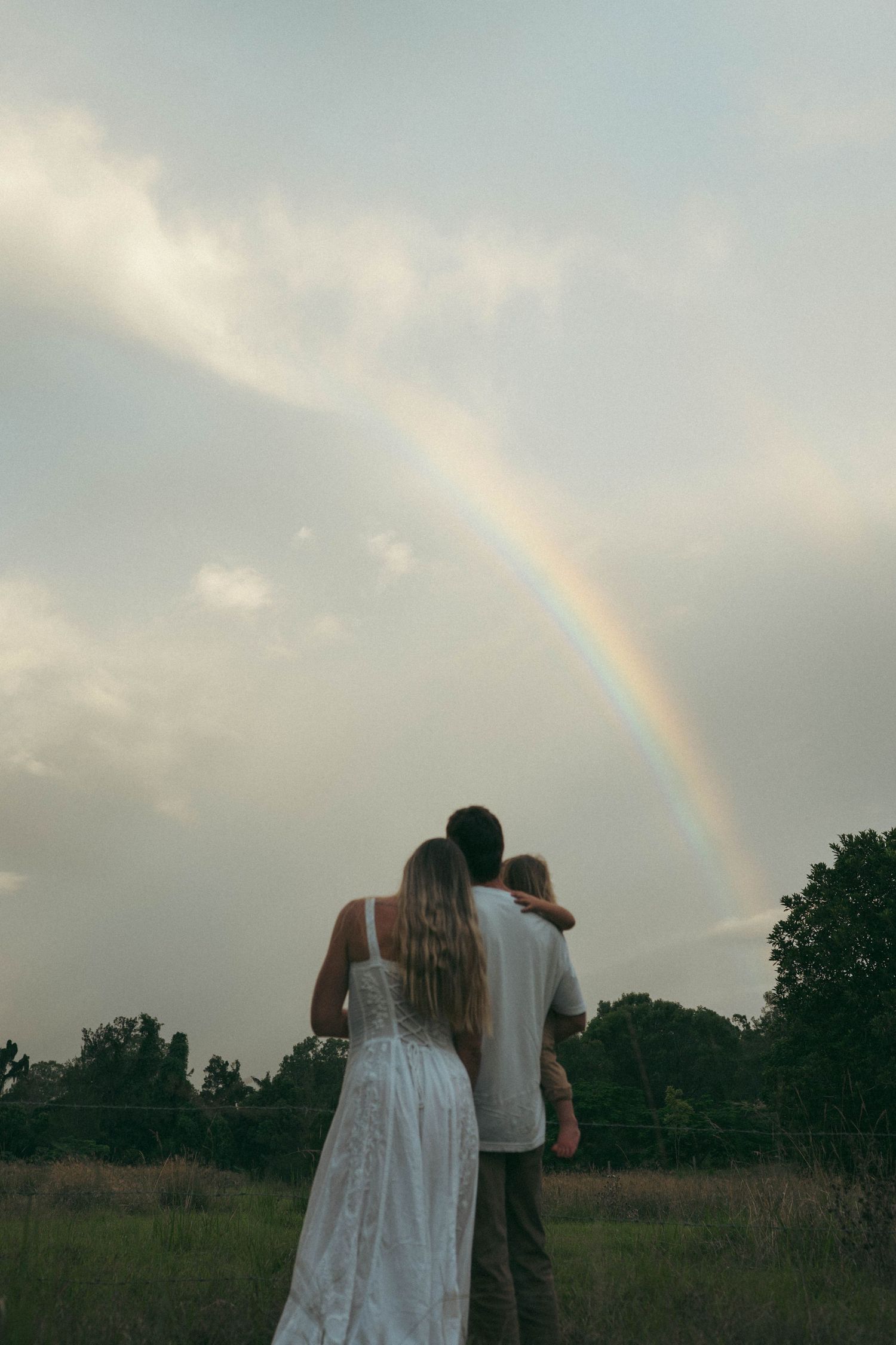 Couple with baby watching rainbow over green field at dusk.