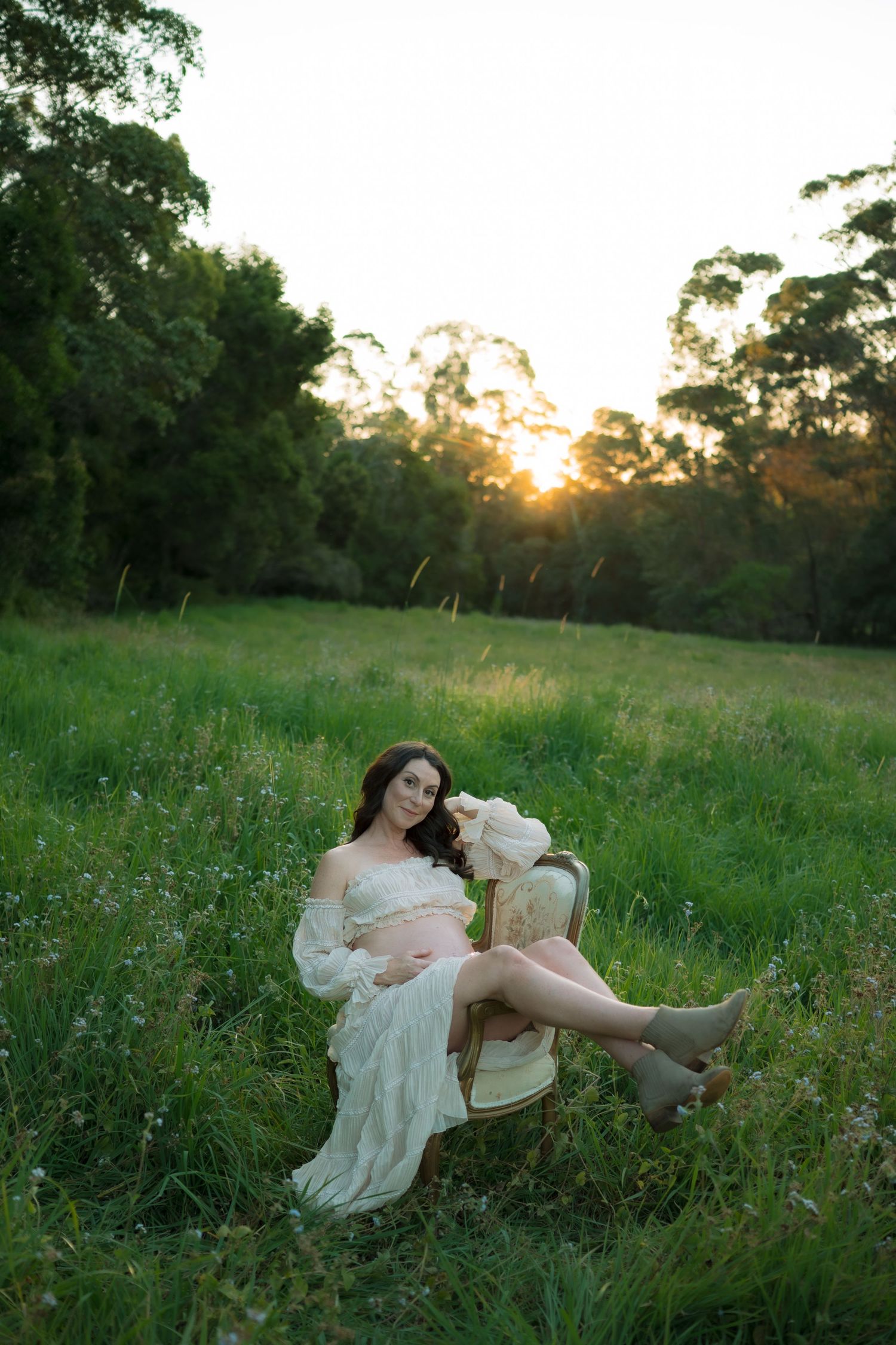 Pregnant woman in flowing white lace dress relaxing on vintage chair in green field at sunset.
