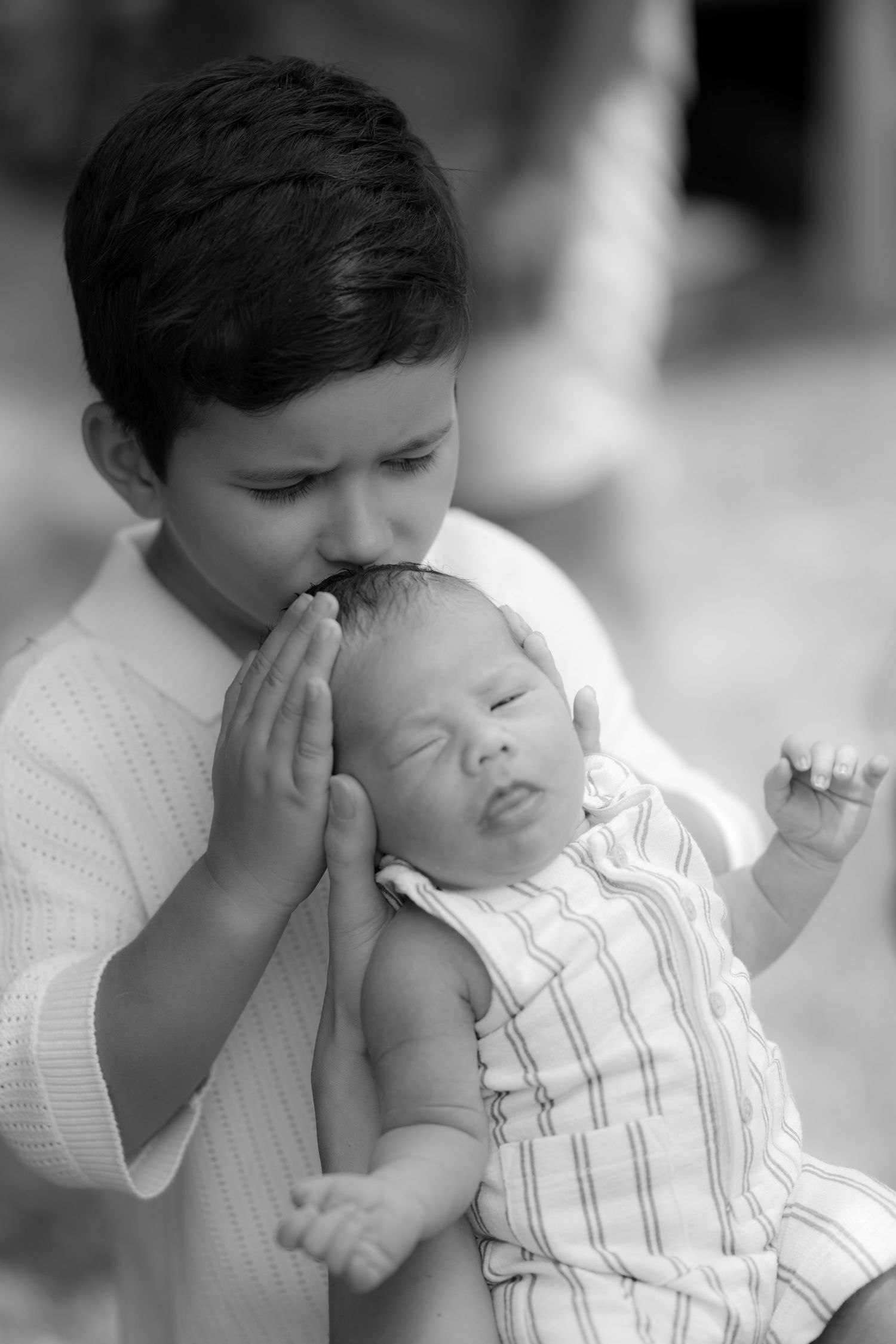 Black and white photo of a young boy tenderly kissing a sleeping newborn baby he holds in his arms.