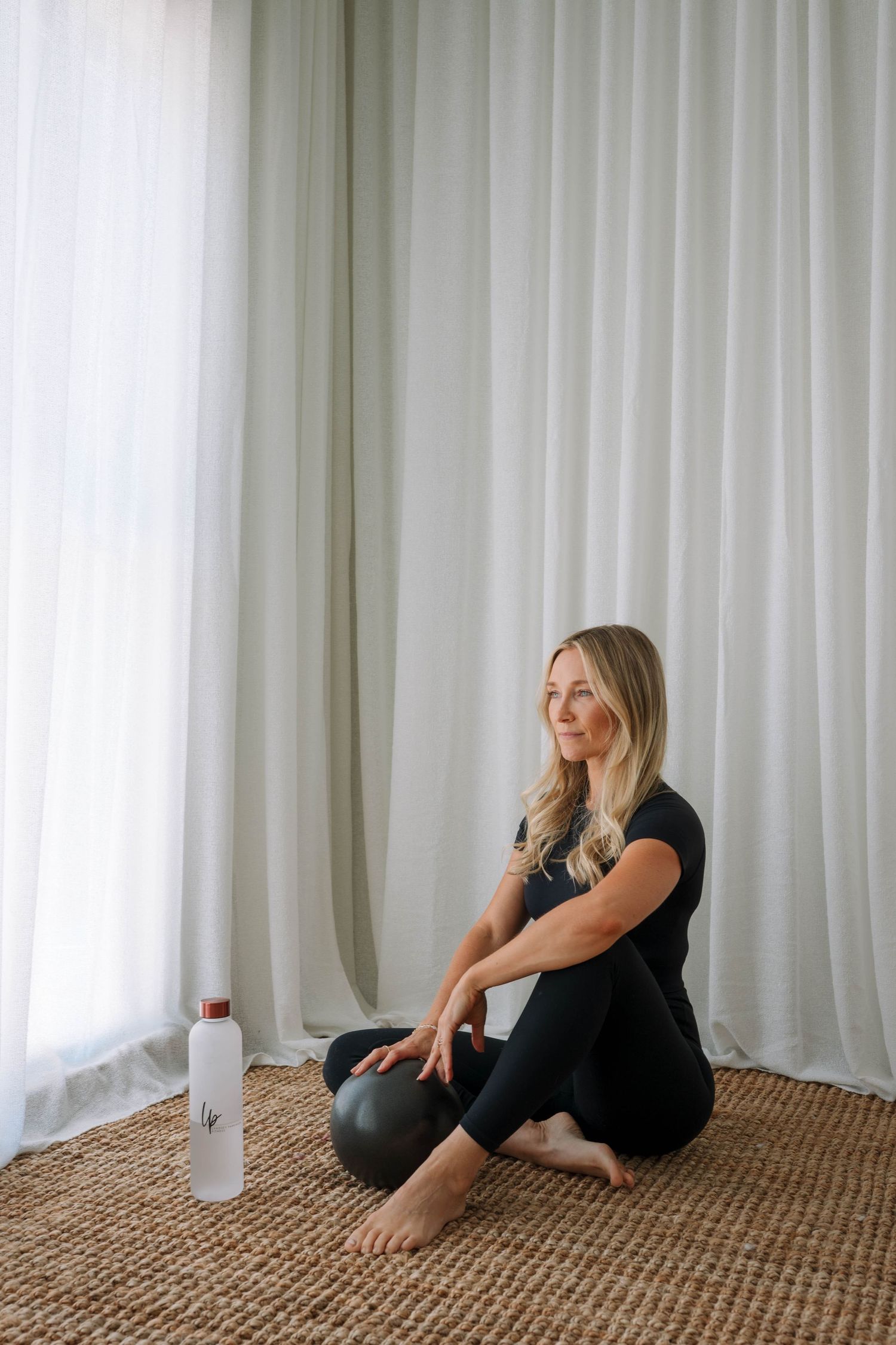 Blonde woman in black activewear sitting on jute rug with pilates ball and water bottle by curtains.