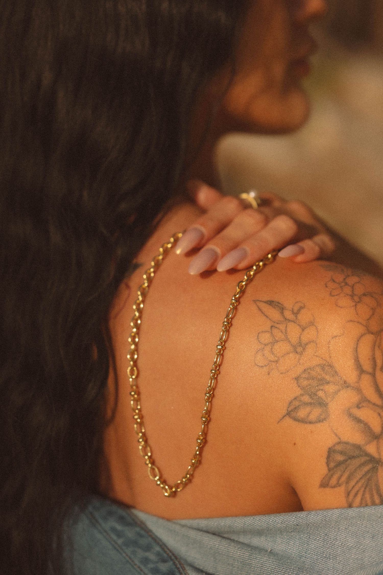 Close-up of gold chain necklace on woman with floral shoulder tattoo in warm light.