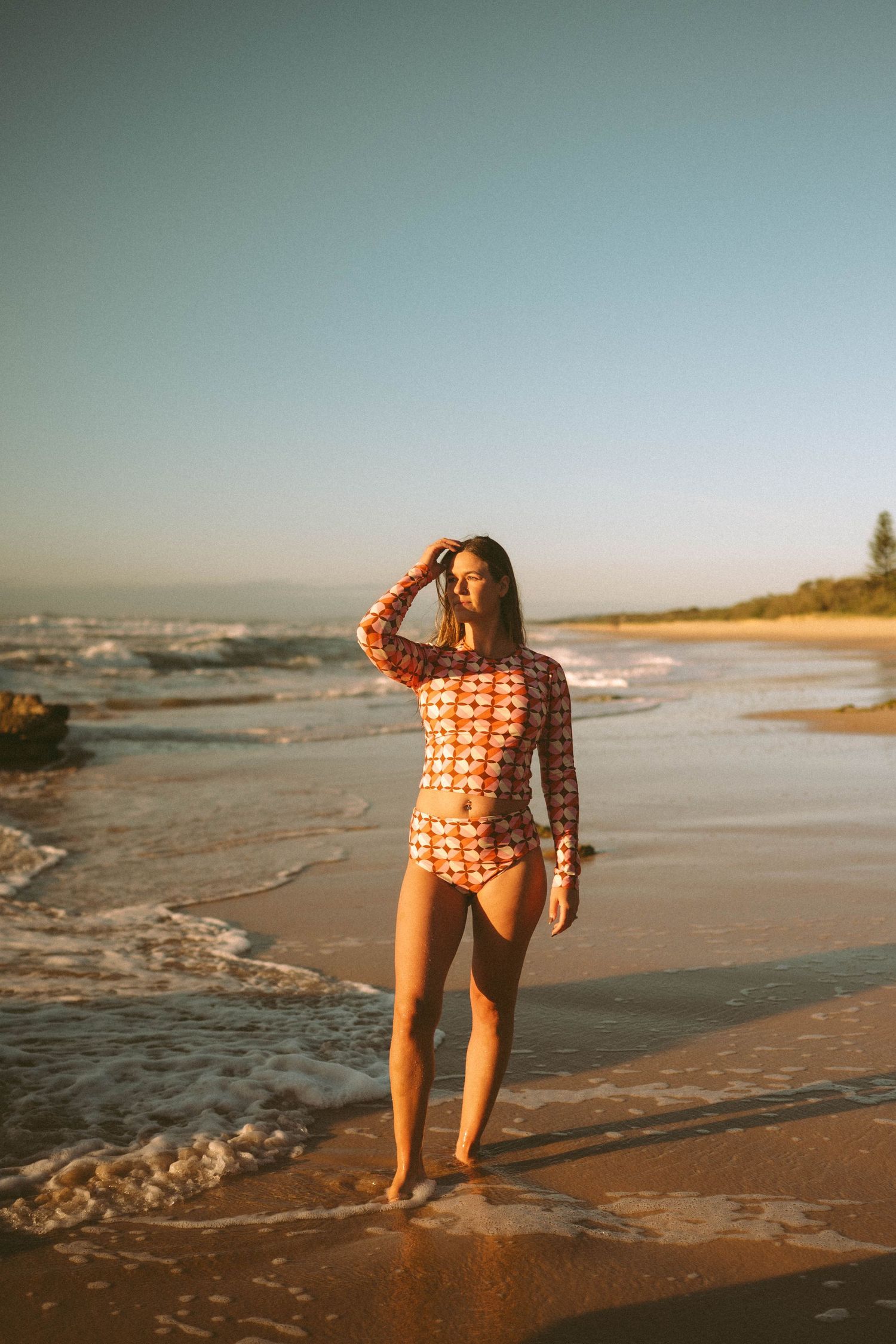 Woman in orange geometric rashguard one-piece swimsuit shielding eyes on sunny sandy beach shoreline.
