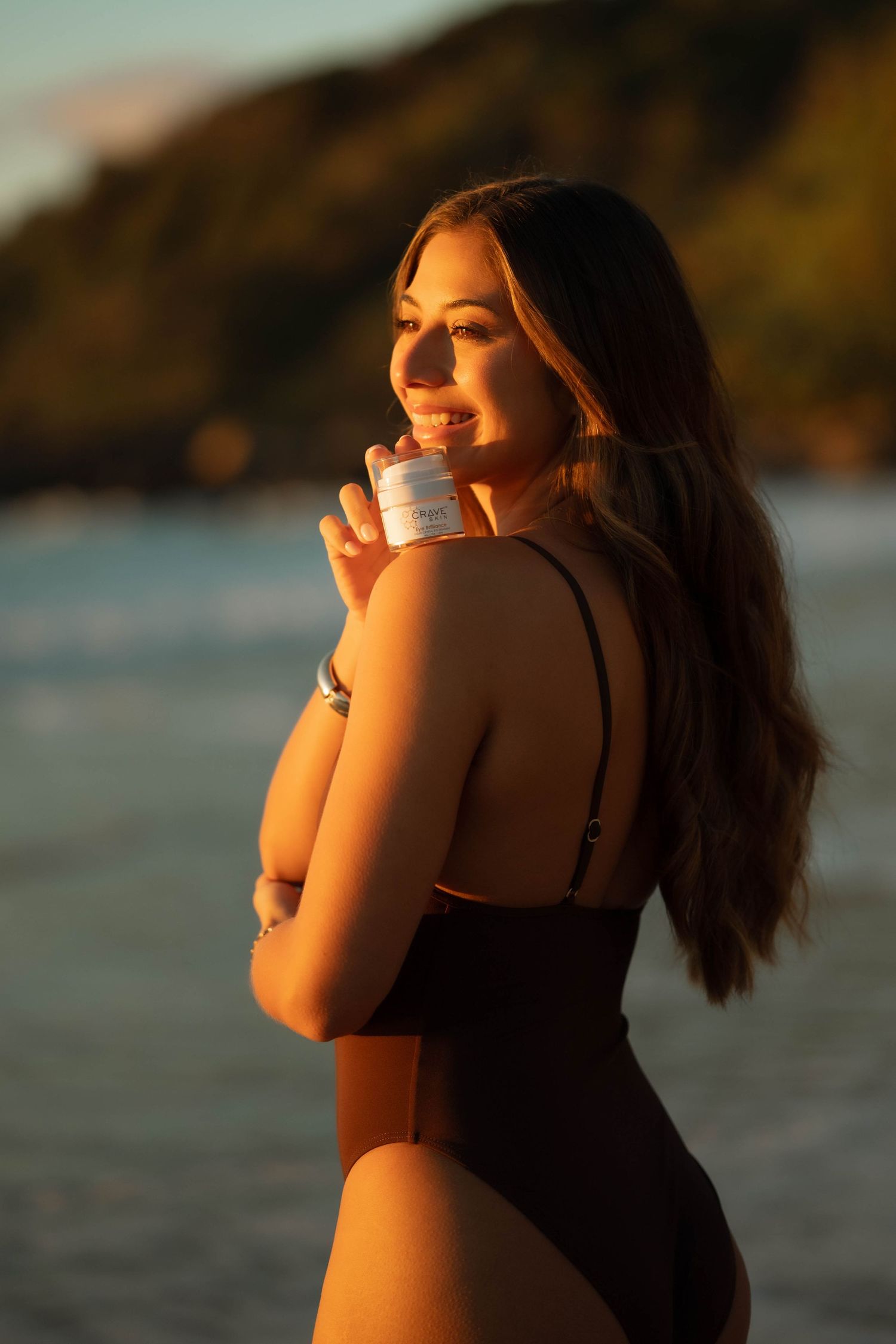 Woman in black swimsuit holding skincare product on tropical beach at golden hour sunset.