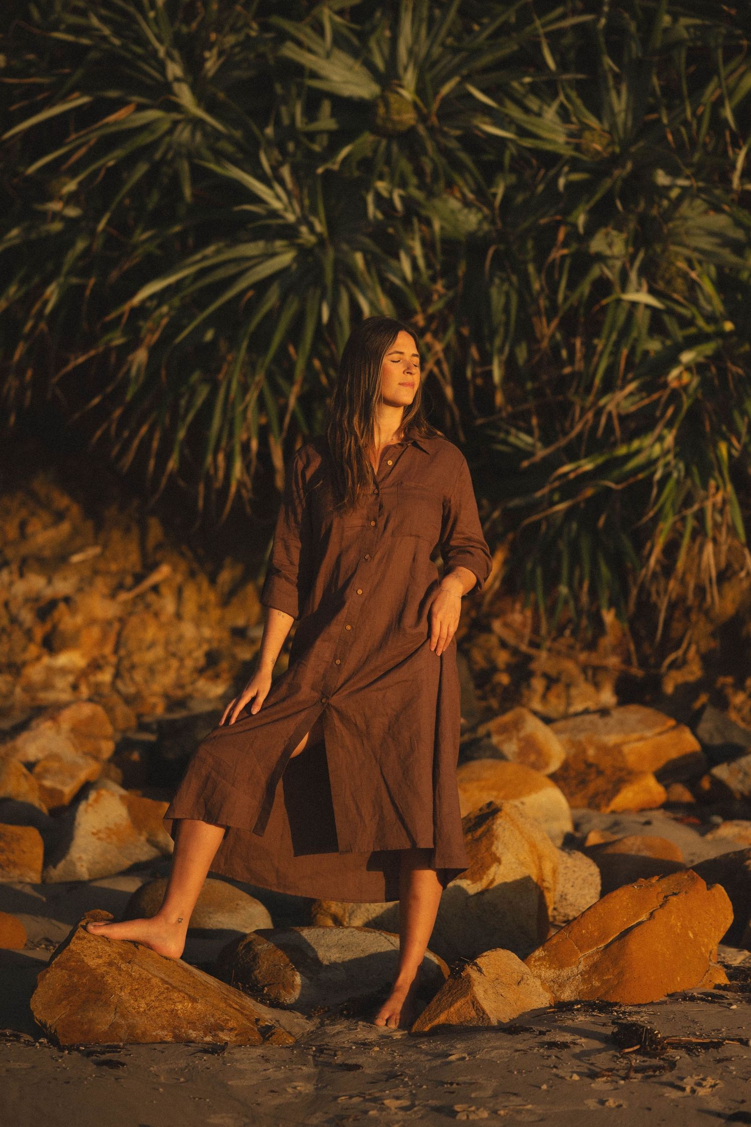 Woman in brown midi dress standing barefoot on rocks near tropical plants in warm golden sunset light.