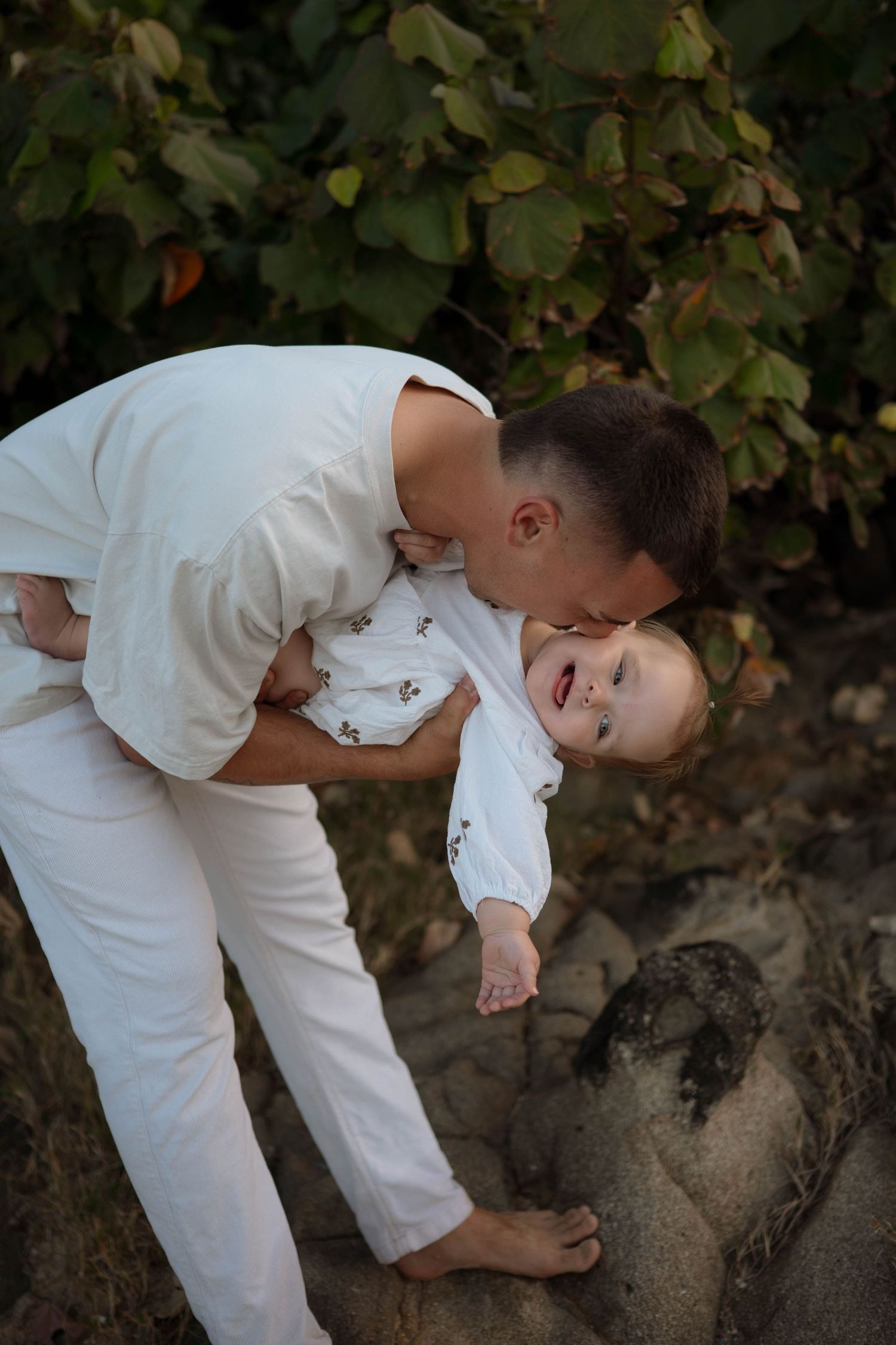A parent in white clothing kisses a laughing baby outdoors near green foliage on a sandy surface.