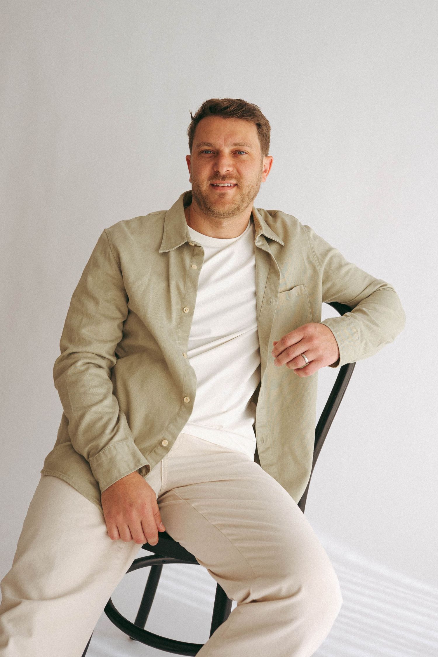 Man in beige shirt and white tee sits relaxed on chair against white background.