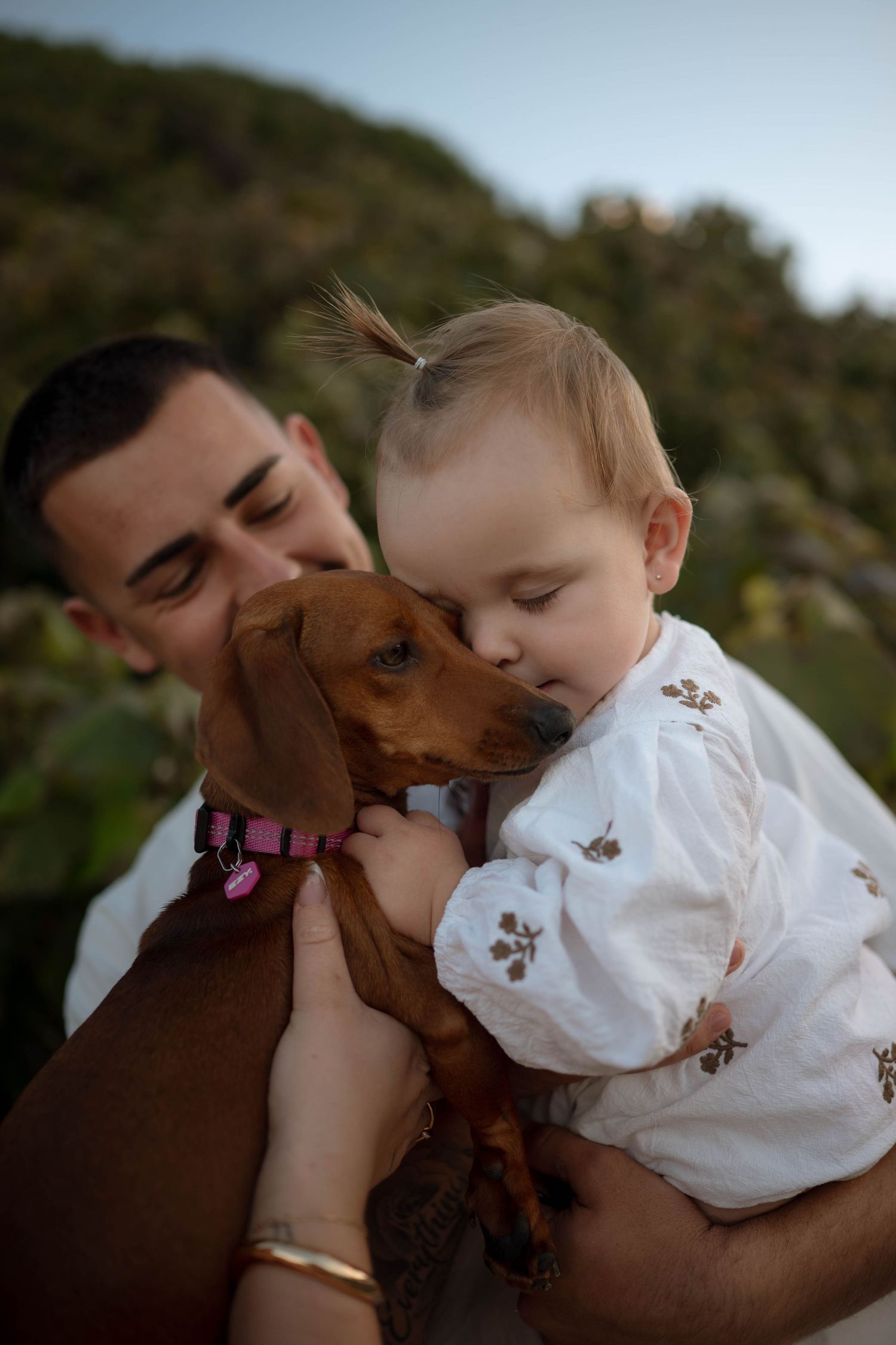 A man holds a baby girl and a dachshund dog outdoors, as the baby gently nuzzles the brown dog.