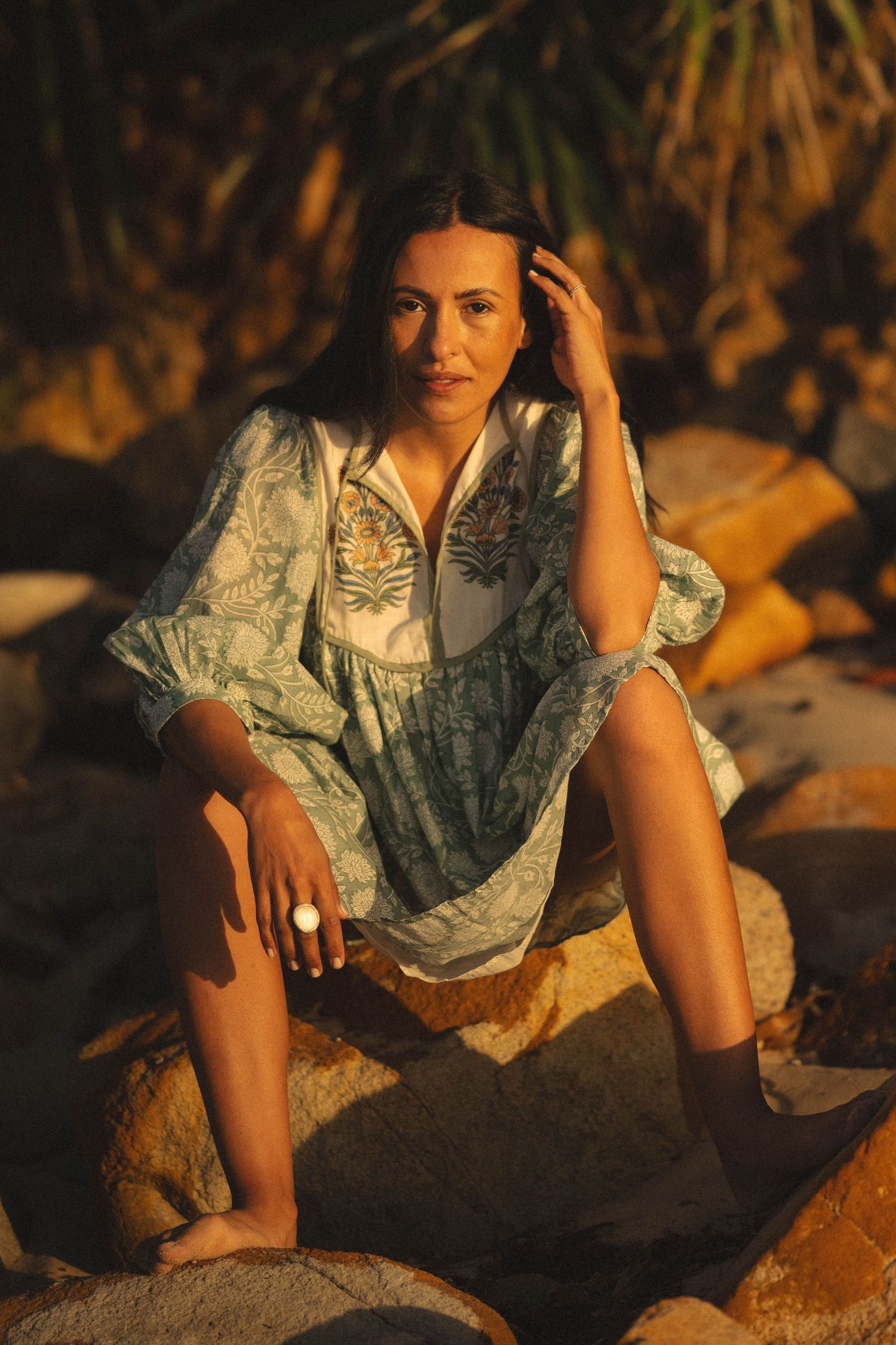 Woman in floral dress sitting on rocks at golden hour sunset with tropical foliage background.