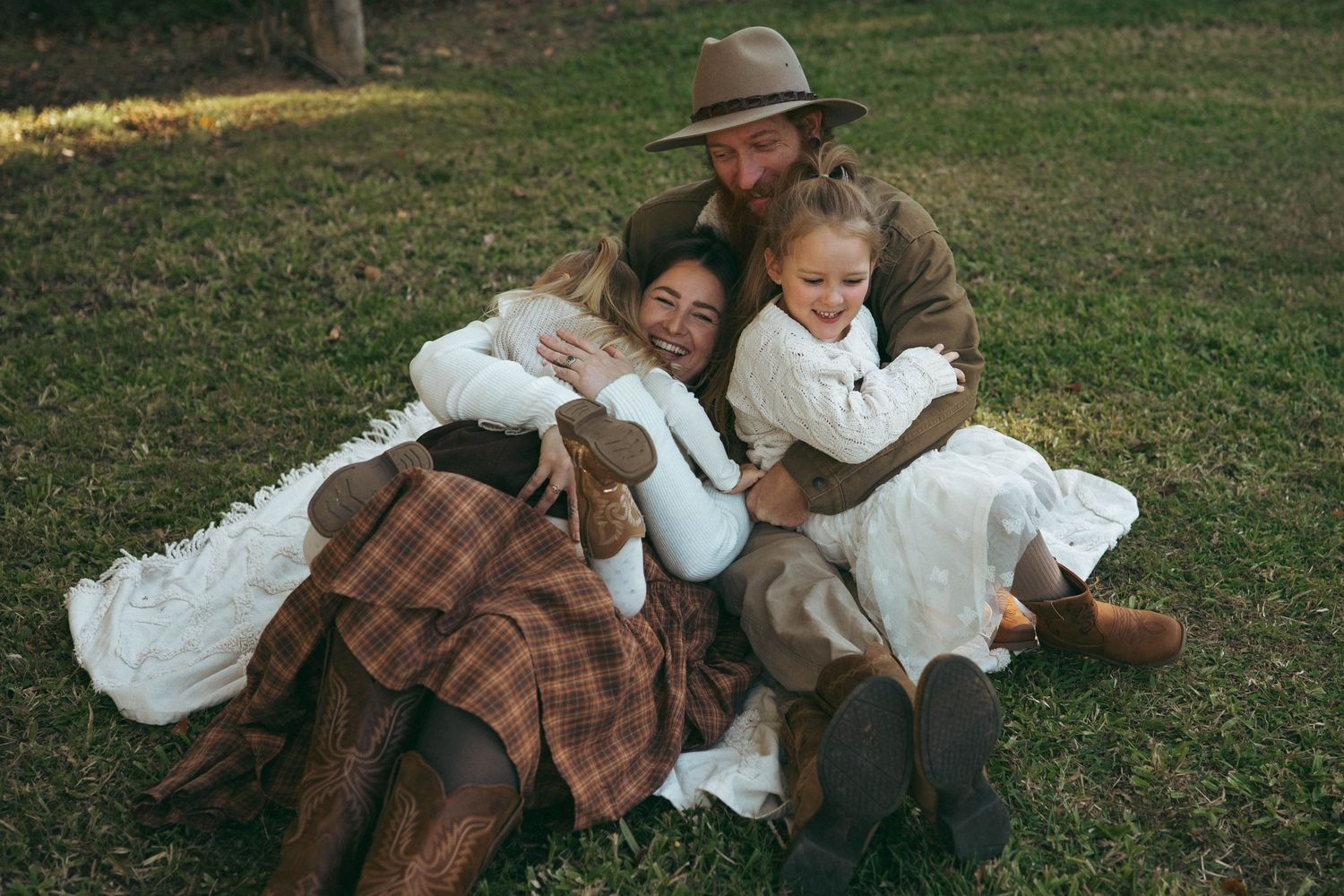 Family of four sharing a warm embrace on grass, dressed in rustic country attire with plaid and white dresses.