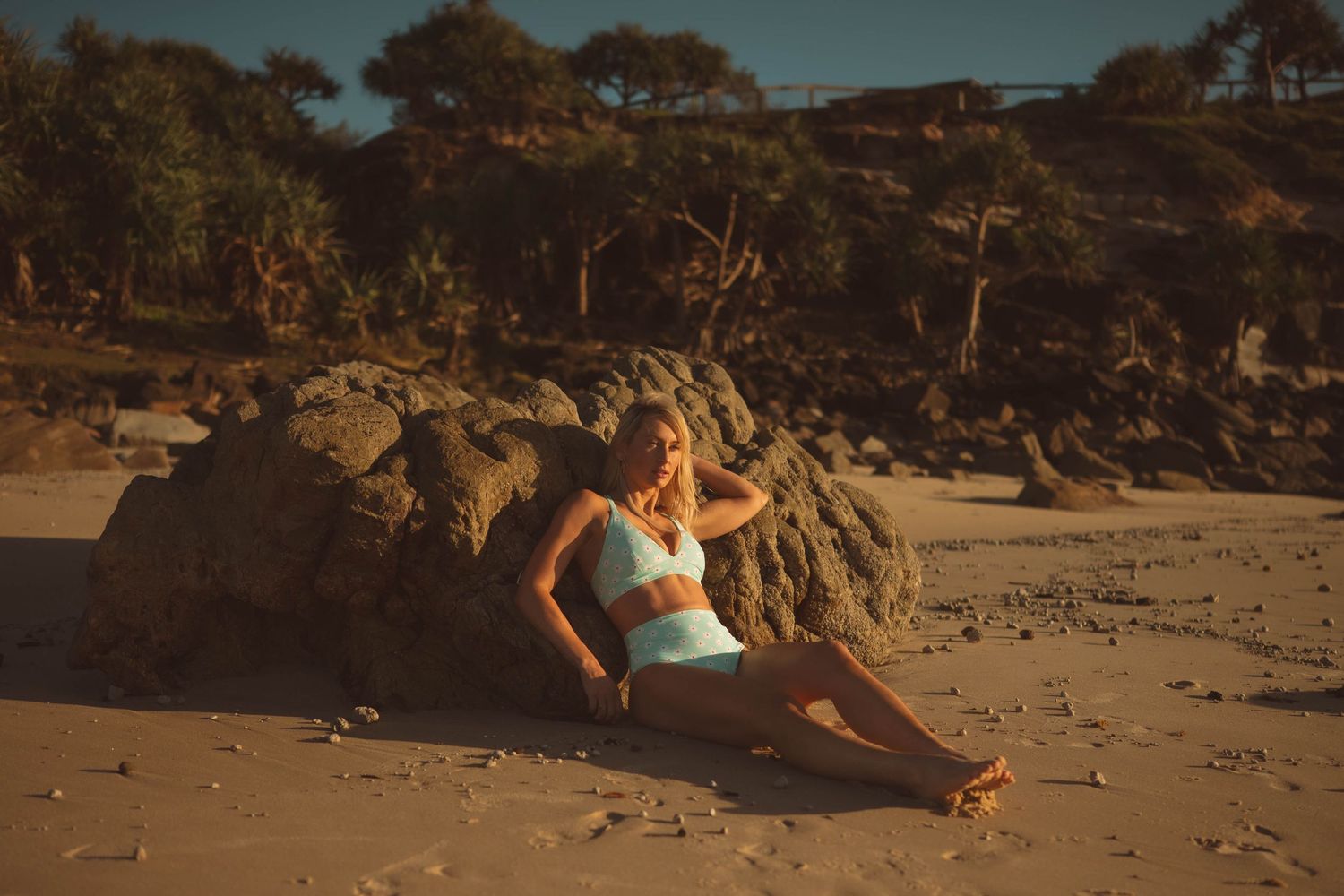 Woman in light blue swimwear relaxing against large rock on sandy beach at sunset.