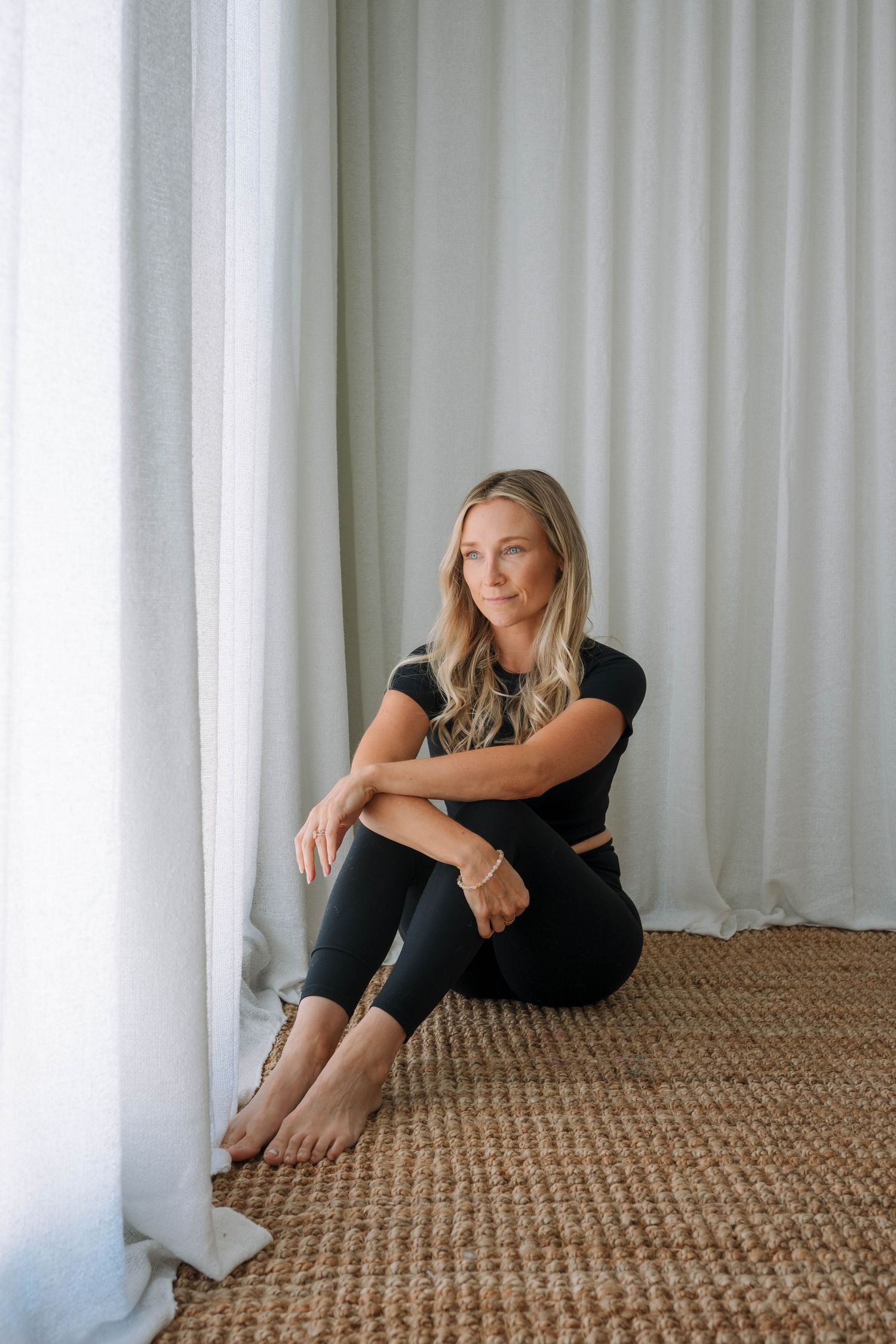 Blonde woman in black crop top and leggings sitting barefoot on jute rug beside sheer curtains.