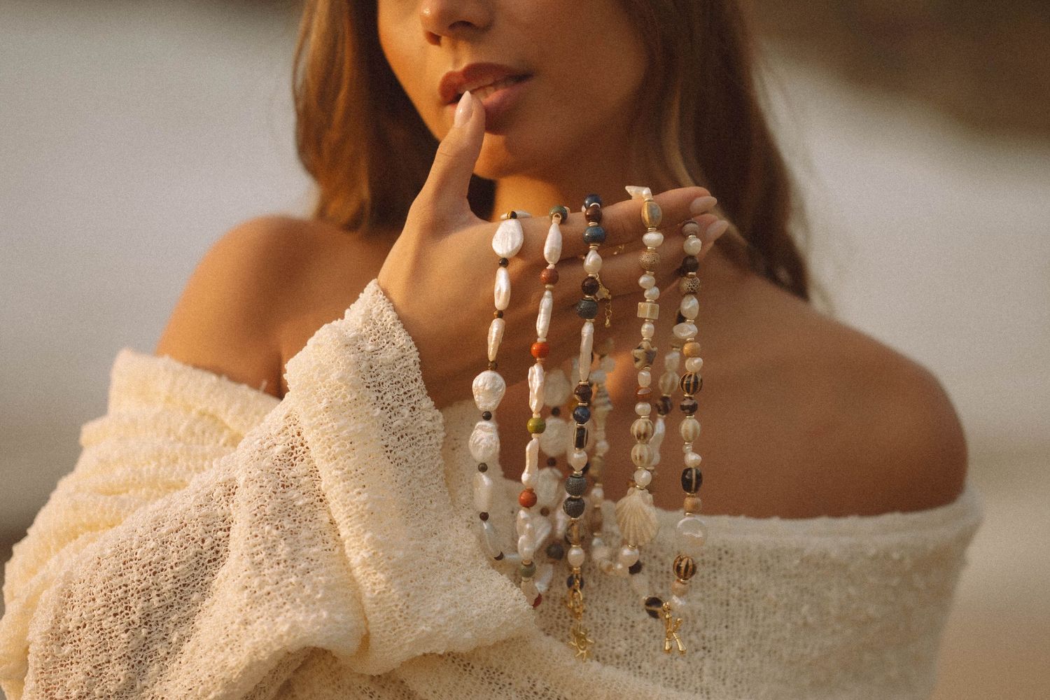 Woman in off-shoulder cream sweater holding multiple pearl and beaded necklaces.