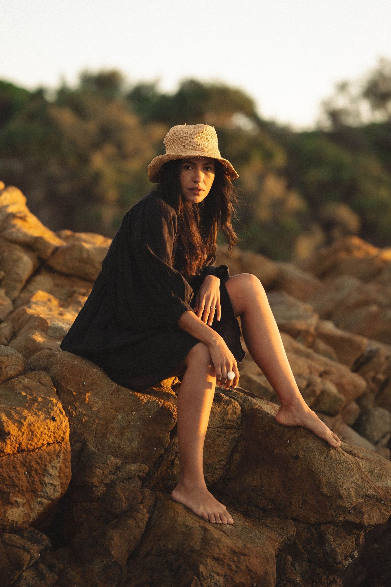 Young woman in black dress and straw hat sitting barefoot on rocks at golden hour sunset.