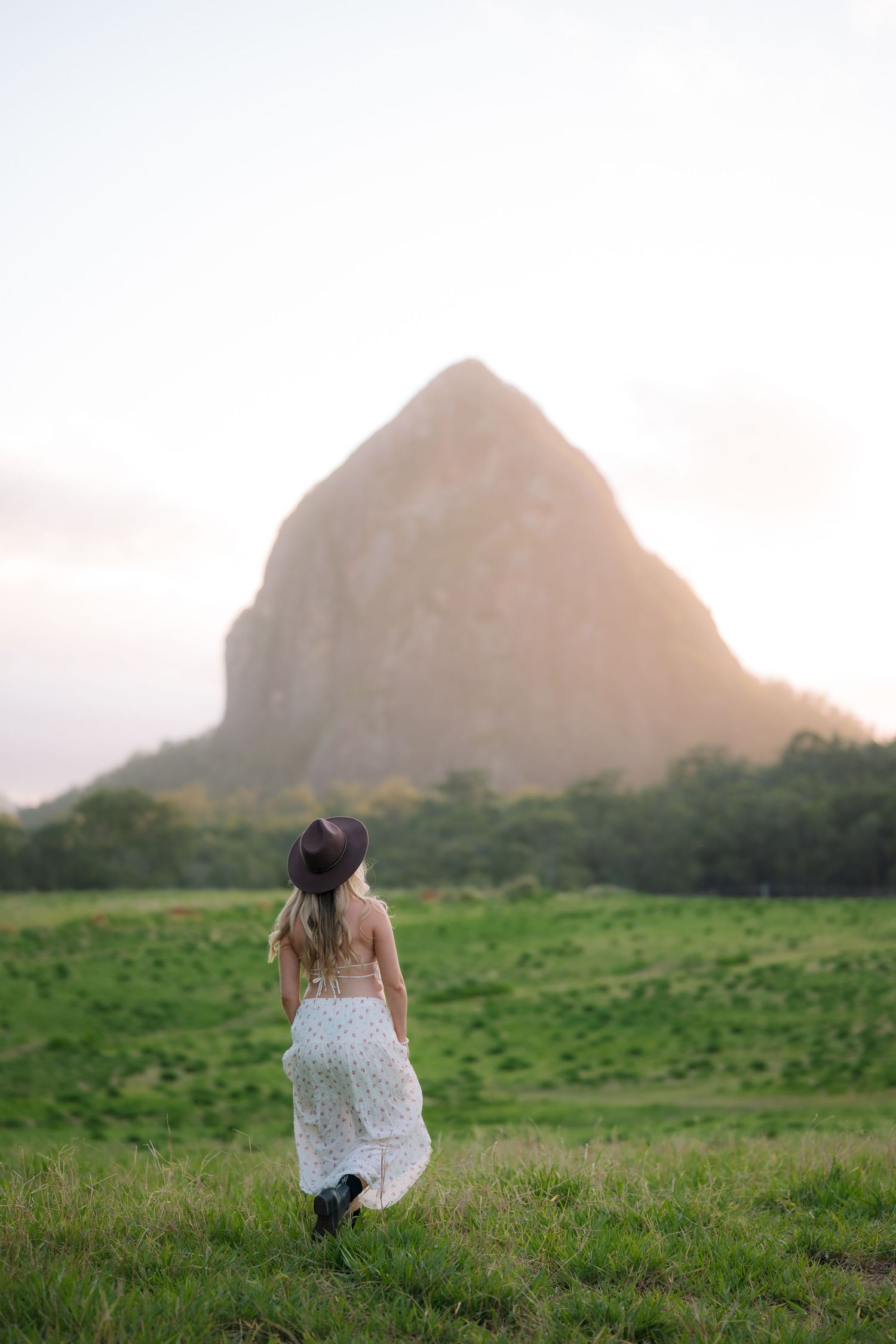 Pregnant woman in white dress and hat walking toward rocky mountain peak at golden hour.