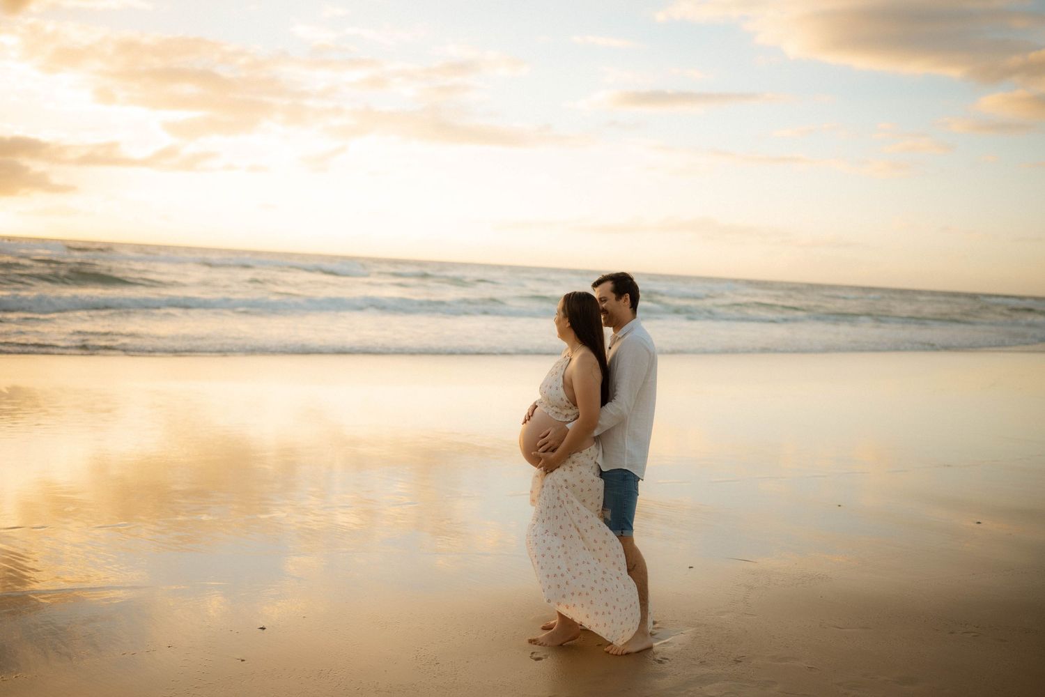 Couple embracing on beach at sunset, pregnant woman in floral dress and partner sharing tender moment.
