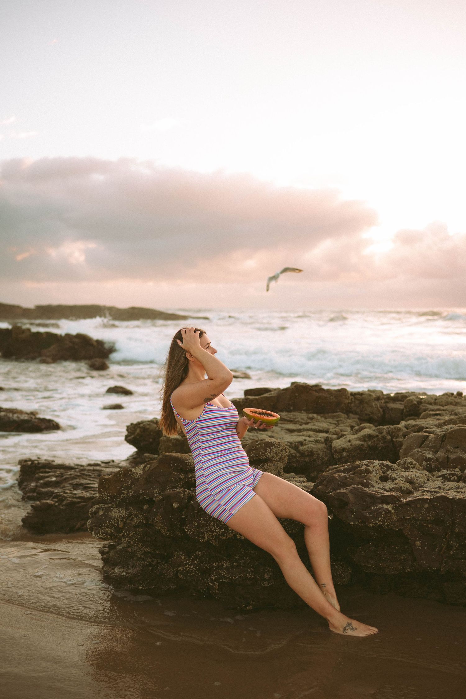 Woman in striped sundress sitting on rocky beach at sunset with a seagull flying overhead.